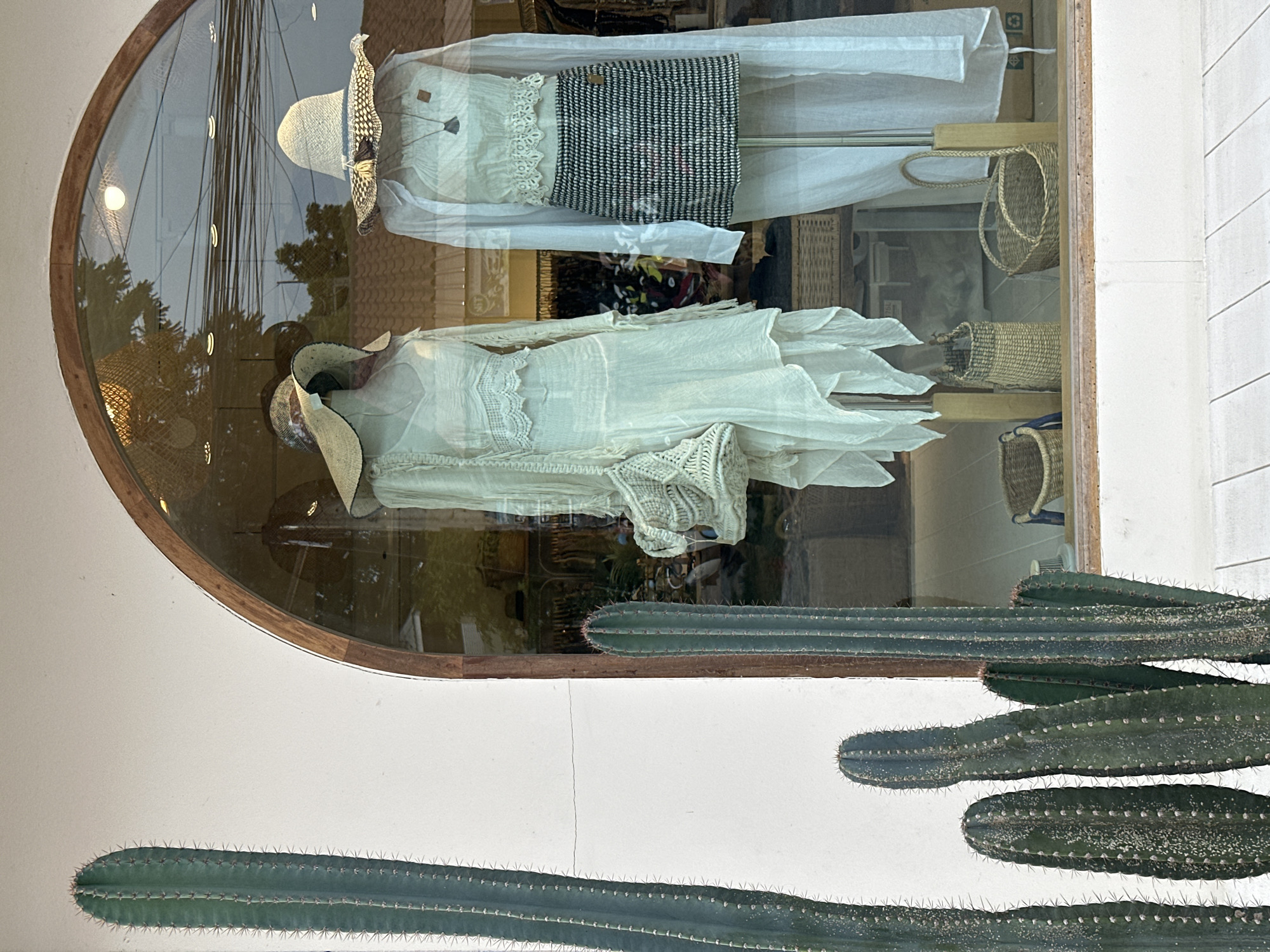 A store window display features two mannequins dressed in light-colored, bohemian-style clothing. The mannequin on the left wears a long, tiered dress with lace accents and a wide-brimmed straw hat. It carries a woven bag. The mannequin on the right is dressed in a white cropped top with lace trim, a black and white patterned skirt, and a straw hat adorned with tassels. A white shirt is draped over it. Several tall, spiky cacti are visible in the foreground outside the window. The store appears to be located in a tropical environment, possibly near the beach given the style of clothing and the presence of woven baskets. The reflection in the window shows trees, power lines, and buildings, suggesting an outdoor commercial area in ตำบลเกาะพะงัน, Thailand.