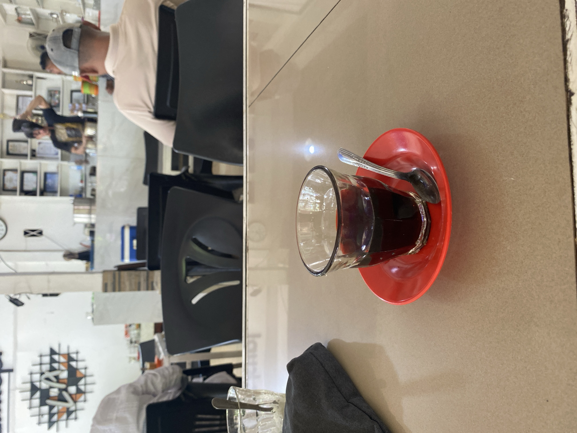A dimly lit indoor establishment, likely a cafe or restaurant, is depicted. In the foreground, a glass of dark liquid, possibly coffee or tea, sits on a red saucer. A spoon rests beside the glass. A dark cloth bag is visible to the left. The background shows blurred figures of patrons and staff. One individual is seen preparing a drink behind a counter, while another person in a grey cap faces away from the camera. The walls are adorned with various framed pictures and shelves stocked with items. The overall ambiance suggests a casual dining or meeting space. The establishment appears to be located in Lhokseumawe, Indonesia, as suggested by the wall art which displays the letters "LA" in a stylized font with tribal geometric patterns, possibly referencing the city or region.