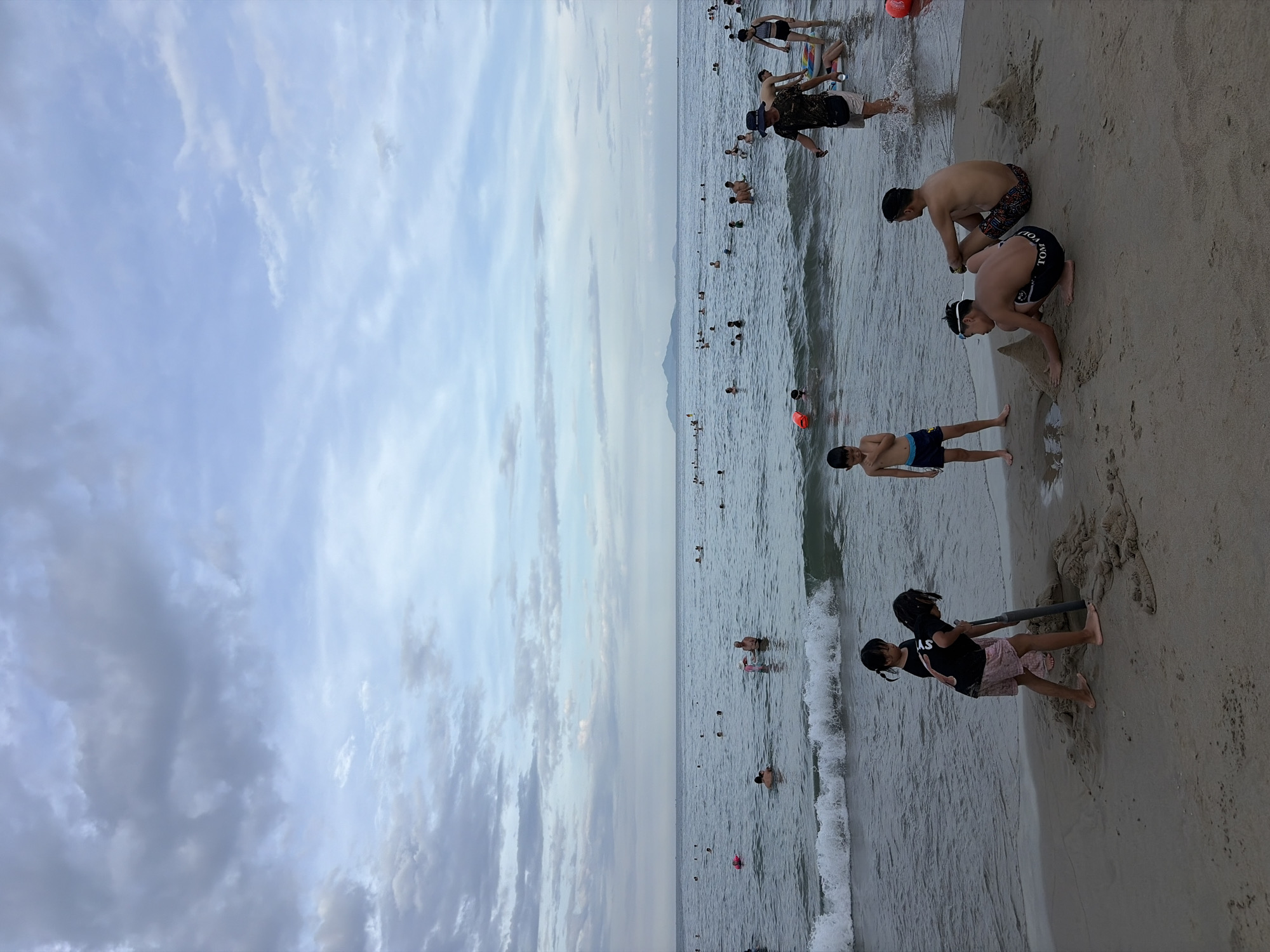 The image depicts a lively beach scene on a cloudy day, likely in Đà Nẵng, Vietnam. The ocean stretches out with numerous people swimming and playing in the water. In the foreground, on the sandy shore, two young girls are engaged in digging in the sand with a shovel. To their right, two boys are also playing in the sand, one constructing a sandcastle and the other sitting nearby. Further in the background, many more people are scattered across the water, some swimming, others wading. The sky is filled with a dramatic display of grey and white clouds, suggesting an overcast or potentially impending weather change. In the far distance, a hazy coastline with mountains can be seen, typical of the Đà Nẵng region. The overall atmosphere is one of leisure and enjoyment, with a strong sense of community and family activity on a popular beach.