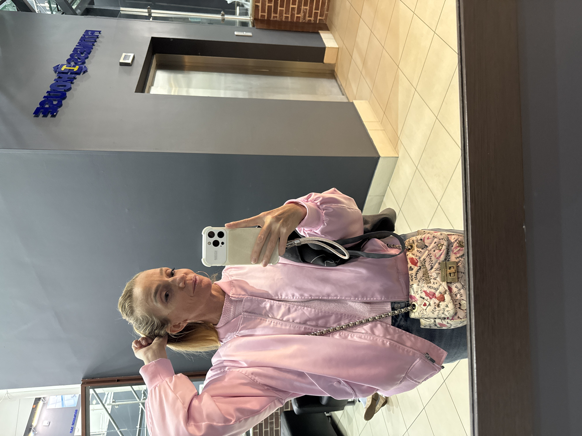 A fair-skinned, mature woman takes a mirror selfie in a room with an elevator and beige tile flooring. The woman wears a pink jacket, a white purse with chain strap, and light-wash jeans. She holds a white cell phone in her right hand. The wall behind her is grey and features the blue letters of “RADIOHOUSE” and an elevator. The scene is in Saint Petersburg, Russia.