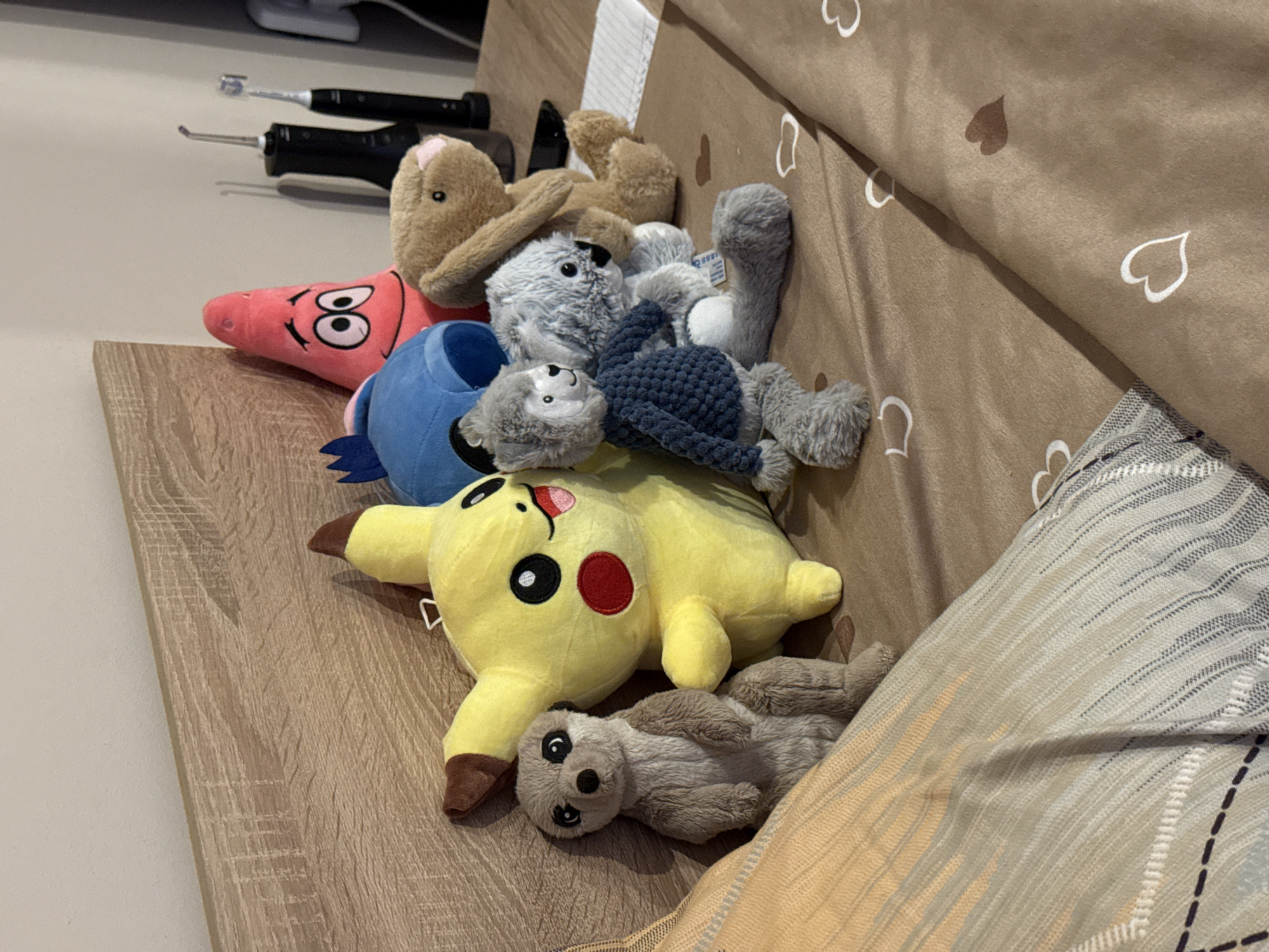 A variety of stuffed animals are piled together on a light brown wooden surface that is adjacent to a bed. The stuffed animals include: Patrick Star (pink), a blue character (possibly Stitch), Pikachu (yellow), a brown bunny, a light-colored monkey, and another gray stuffed animal. Behind the toys, there are two dental cleaning devices (or similar objects). The bed has a light brown patterned blanket with hearts, and a gray and tan striped pillow partially covered by a beige and yellow blanket. The location cannot be determined to be in Lovech, Bulgaria, based on the given image.