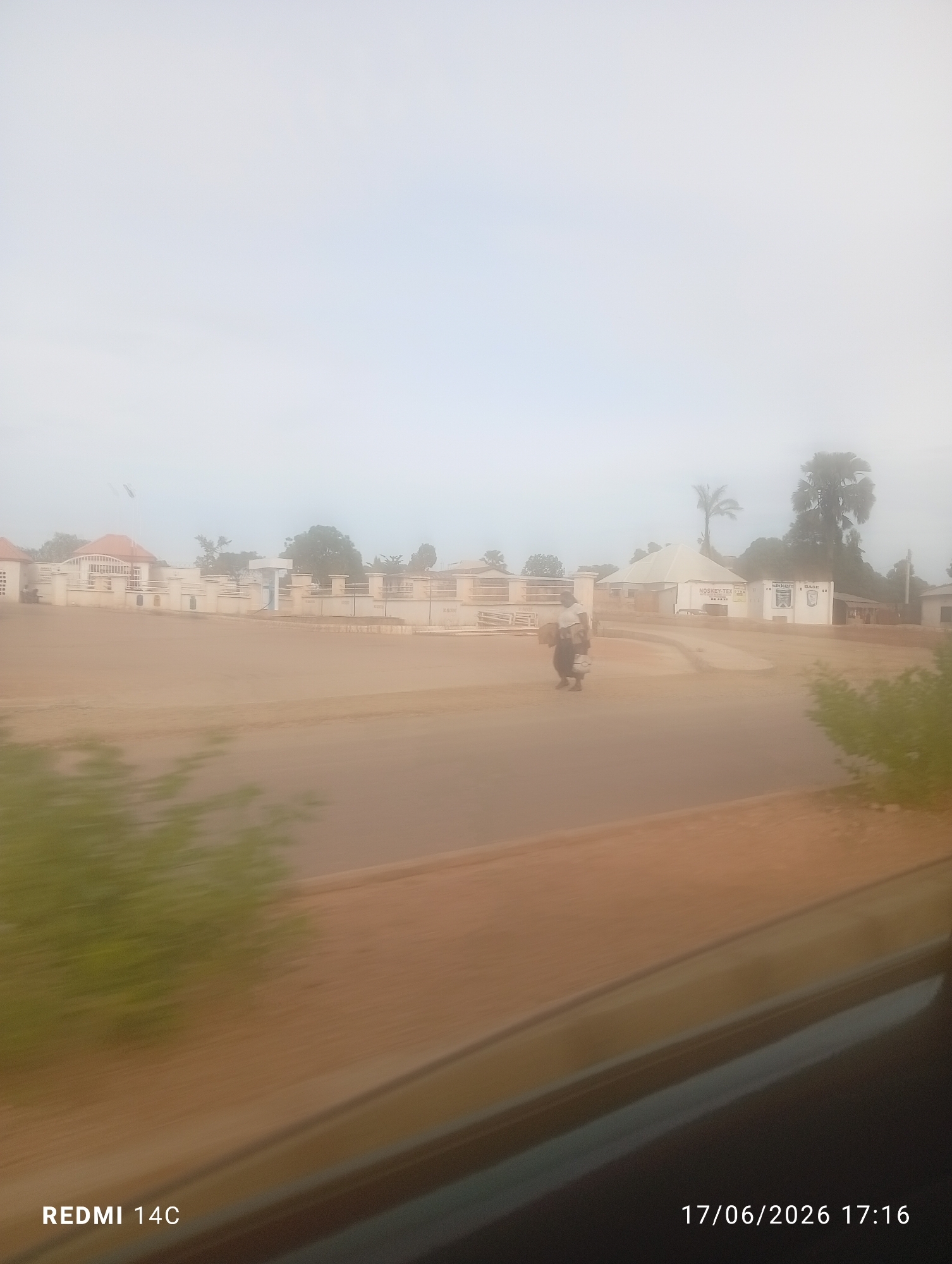 The image depicts a street scene in Bukuru, Nigeria, on a hazy day. In the foreground, a road is visible with blurred greenery on the left. Further back, a building complex with beige walls and red-tiled roofs is present. A white fence runs along the front of the complex. To the right of the complex, a white structure with a light-colored conical roof stands, with signage that reads "NOSKEY-TEX GLOBAL" and "SIKKENS". Two palm trees are visible behind this structure. A person is seen walking on the road, carrying bags. In the middle ground, a motorcycle with two riders is moving. The sky is overcast and hazy. The bottom right corner of the image displays a timestamp: "17/06/2026 17:16", and the bottom left corner displays "REDMI 14C".