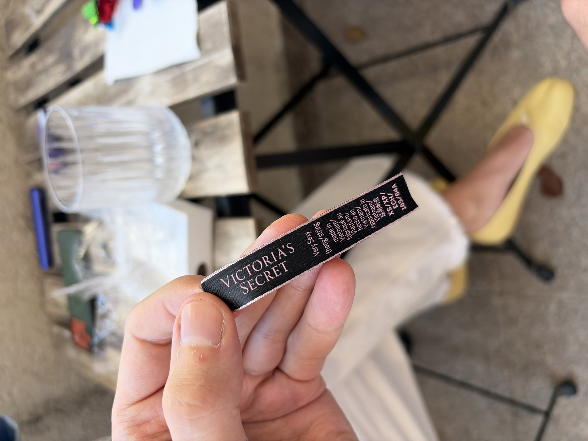 A close-up, high-angle shot captures a person's right hand holding a black Victoria's Secret clothing tag. The tag prominently displays "VICTORIA'S SECRET" in white lettering. Further text on the tag, partially visible, includes "Very Sexy", "thong/string", and "made in Vietnam" repeated in multiple languages (French, Spanish, Italian, and Vietnamese characters), indicating the garment's origin. Size details like "XS/XP/ ECH/" and "165/64A" are also legible. The hand shows clear fingernails on the thumb and index finger.
The background is intentionally blurred, suggesting a daytime setting in Budva, Montenegro. Visible elements include a light-colored wooden slatted surface, likely a table, and a clear plastic cup resting on it. Parts of a dark metal frame, possibly belonging to a chair, are also seen. A blurry yellow shoe or sandal on a foot is visible in the lower right, implying another person or the wearer of the tag is present in the scene. The overall impression is casual and domestic.