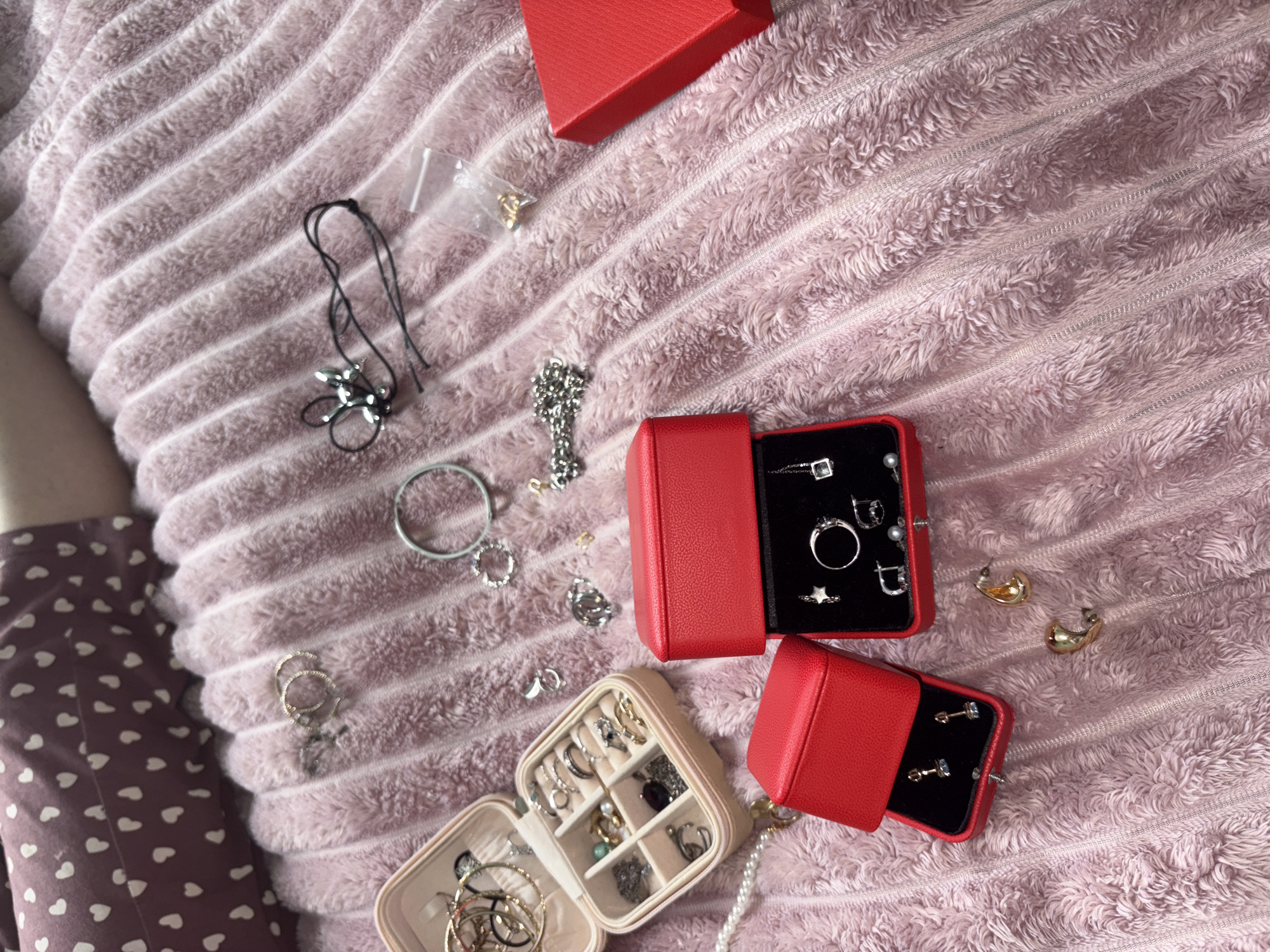 An overhead, medium shot captures a collection of jewelry arranged on a plush, pink, ribbed blanket. The blanket takes up the majority of the frame, with a portion of a purple, heart-patterned garment visible in the upper left corner, suggesting a person is partially in the frame, though no body parts are visible.
Scattered across the blanket are various jewelry items and storage containers. Two open, beige, compartmentalized jewelry boxes are prominent on the left. The larger one, on top, contains numerous rings and bracelets. The smaller one, below it, holds rings, earrings, and other small trinkets.
To the right of the jewelry boxes are two bright red, lidded jewelry boxes, one stacked partially on top of the other. The upper red box is closed, while the lower one is open, revealing a black interior with several rings and earrings. A separate, open red jewelry box sits to the left of the stacked ones, displaying a pair of earrings.
Additional jewelry items are dispersed on the blanket, including a delicate silver chain, a grey beaded bracelet, several pairs of earrings, rings, and a single pearl necklace. A small, clear plastic bag containing tiny gold-colored pieces is also visible. The overall impression is one of organized disarray, with the jewelry laid out as if being sorted or displayed. The lighting is soft, casting gentle shadows and highlighting the textures of the blanket and jewelry.
