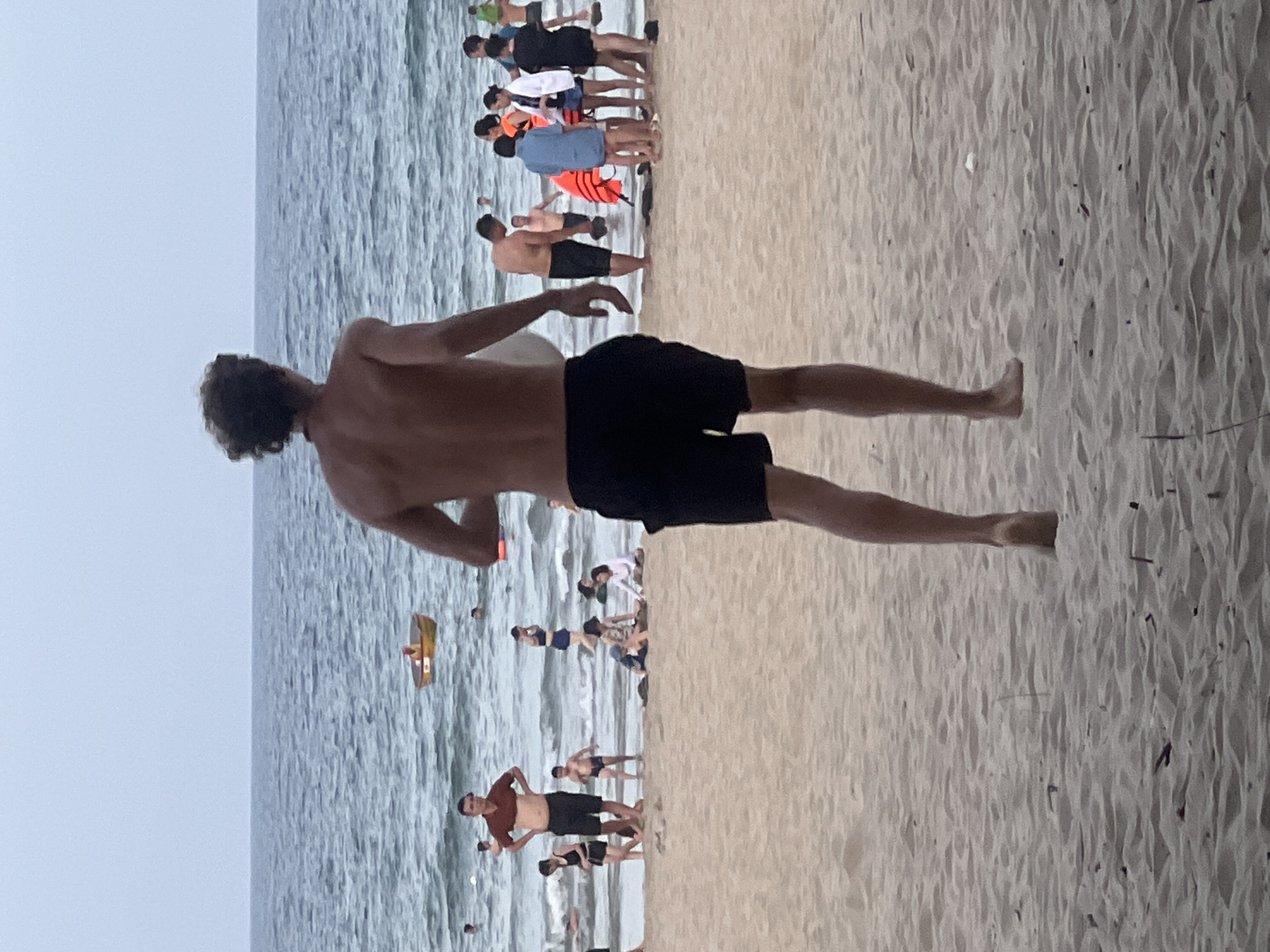 The media file depicts a crowded beach scene in Đà Nẵng, Vietnam. The main subject is a young man in the foreground, shirtless and wearing black swim trunks, standing with his back to the camera and looking out at the ocean. His arms are extended, and he appears to be in motion, possibly mid-stride or stretching.
The background is filled with numerous people on the sandy beach and in the water. The ocean is calm with gentle waves. Several individuals are standing near the water's edge, some wearing life vests in bright orange and blue, suggesting they might be engaged in water activities or are part of a supervised group. Others are further back on the sand, walking or standing in groups. In the distance, a yellow floating object, possibly a small boat or a float, is visible in the water.
The lighting suggests it is daytime, likely late afternoon or early evening, given the soft light and overcast sky. The weather appears to be warm and humid, typical for a beach location in Vietnam. The overall mood is one of leisure and recreation, with people enjoying a day at the beach. There is no visible text in the image.