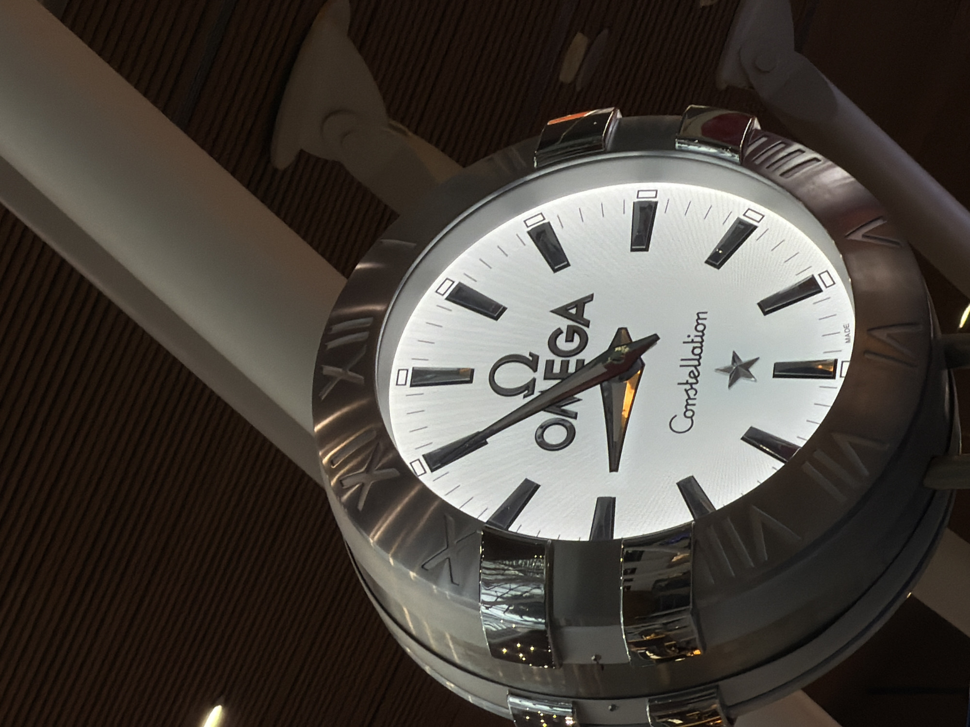 An Omega Constellation clock is displayed in an indoor environment, likely an airport terminal, given the surrounding architecture. The clock face is white with black hour markers, and the Omega logo and "Constellation" text are visible. The clock has Roman numerals etched around its metallic casing. The environment features wooden-paneled ceilings and white structural elements. No people are visible in the image. The clock appears to be a decorative or functional piece within a public space.