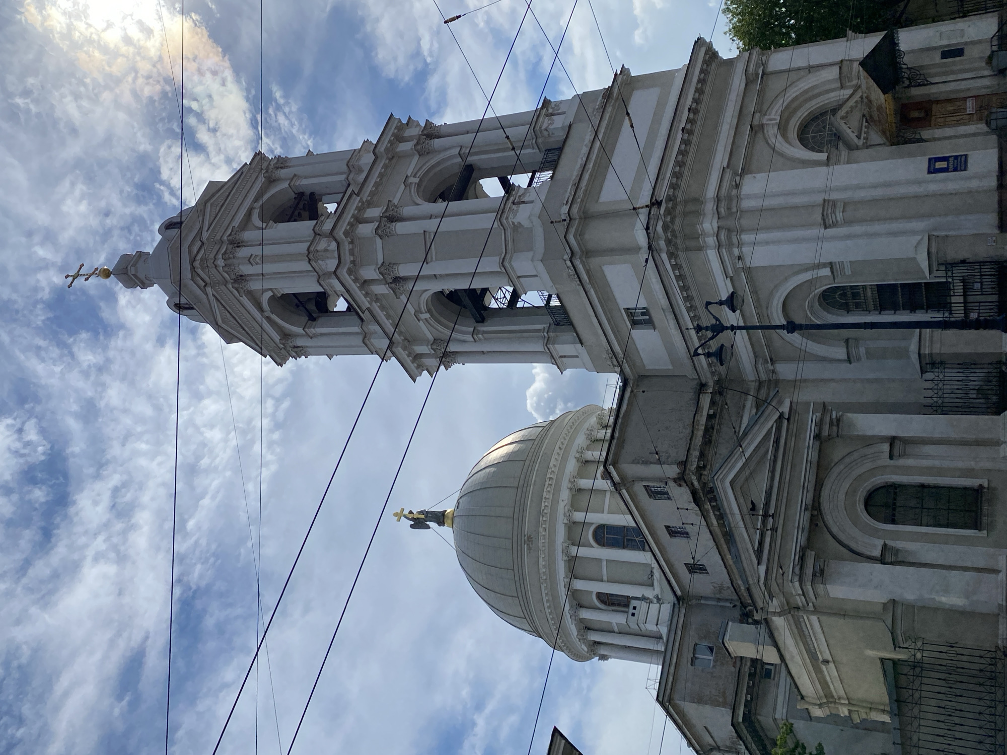 The image depicts a tall, ornate bell tower and a dome of a church, likely in Saint Petersburg, Russia, given the architectural style. The bell tower is square at its base and tapers upwards, featuring multiple levels with arched openings revealing bells within. It is topped with a cross. The dome, a prominent feature of the church, is rounded and features classical architectural elements, including columns and arches. A gilded angel figure holding a cross stands atop the dome. The sky above is partly cloudy, with sunlight breaking through in some areas, creating a dramatic effect. Several overhead wires, characteristic of urban environments in Russia, crisscross the frame, partially obscuring the view of the buildings and sky. The church itself appears to be constructed of light-colored stone or stucco, with intricate detailing. In the foreground, elements of the church's facade are visible, including arched windows and decorative moldings. A black wrought-iron fence and a street lamp add to the urban streetscape.