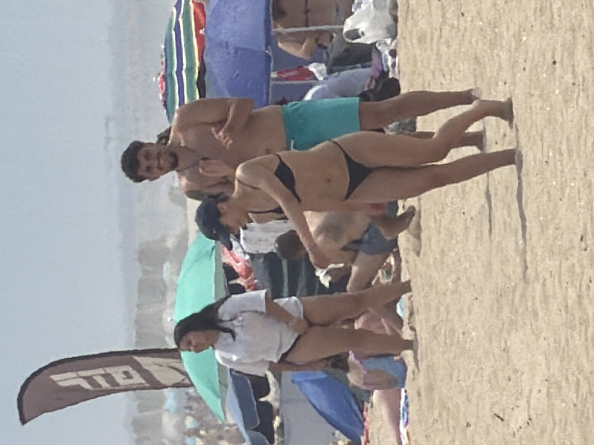 The image depicts a crowded beach scene at Espinho, Portugal, under a bright sky. In the foreground, a woman in a black bikini walks on the sand, her body angled towards the right side of the frame. Her dark hair is pulled back, and she is wearing a dark baseball cap. Behind her and to her right, a shirtless man with dark hair and a beard smiles and claps his hands, wearing turquoise swim trunks. To the left of the woman in the bikini, a woman in a white t-shirt and black bikini bottoms stands looking to the left. A large, dark flag with white lettering is positioned to the left of the women. Beach umbrellas, some colorful and some blue, are visible in the background, along with other beachgoers and the ocean. The sand is a light brown color.