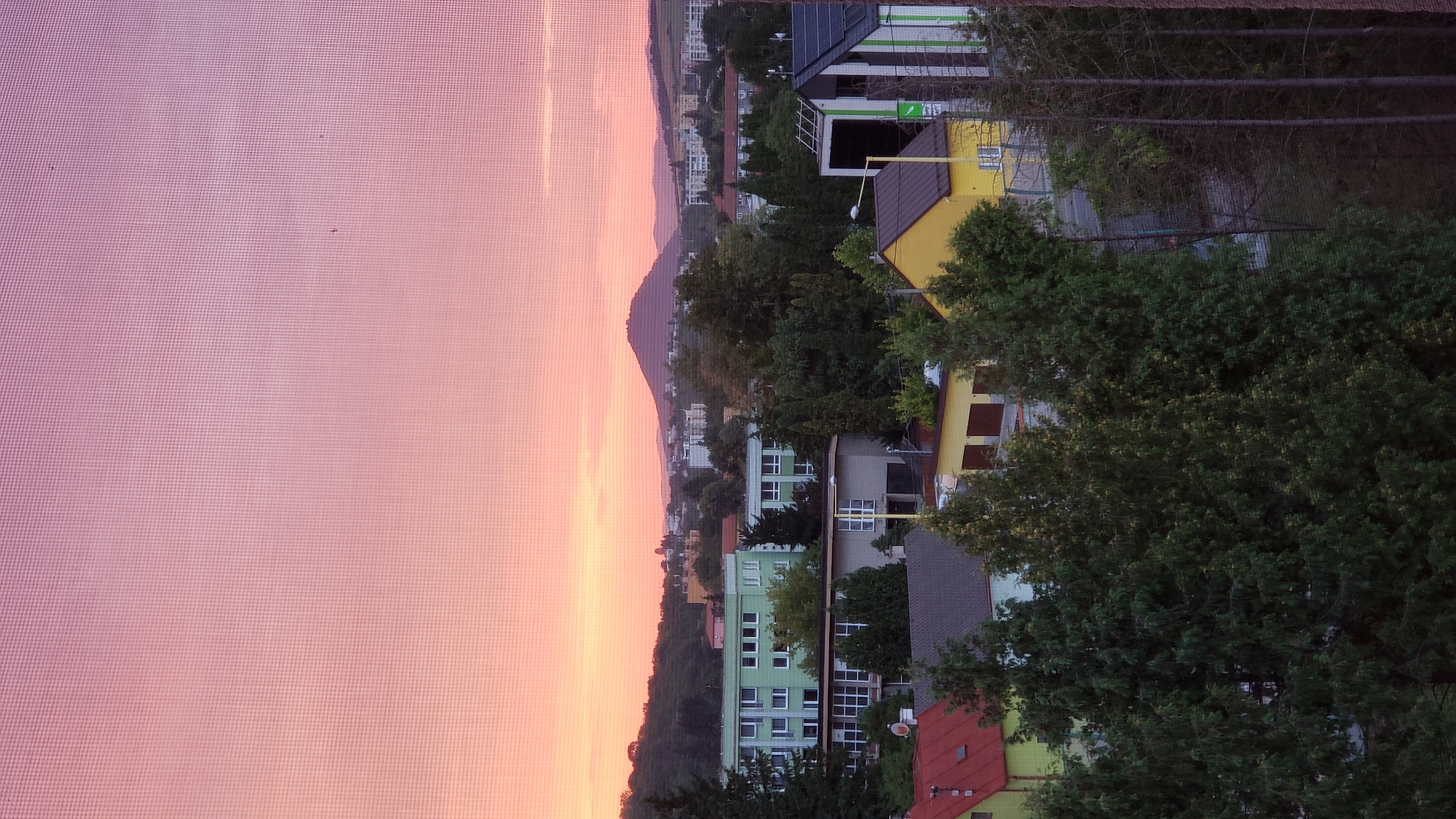 The image captures a panoramic view of a city at sunset. The sky is a gradient of pink and orange hues, with a textured overlay suggesting a screen or mesh. In the mid-ground, several buildings are visible, including a multi-story green building, a lighter colored building, and a gray structure. Trees and lush greenery are interspersed throughout the cityscape. In the background, a conical hill dominates the horizon, with more buildings and distant hills fading into the twilight. The foreground is occupied by dark green trees, obscuring parts of the lower buildings. Based on the characteristic conical hill in the background, this scene is likely depicting the view of the city of Prešov, Slovakia, with the prominent landmark being the Zelená hora (Green Mountain) or a similar prominent geological formation in the vicinity.