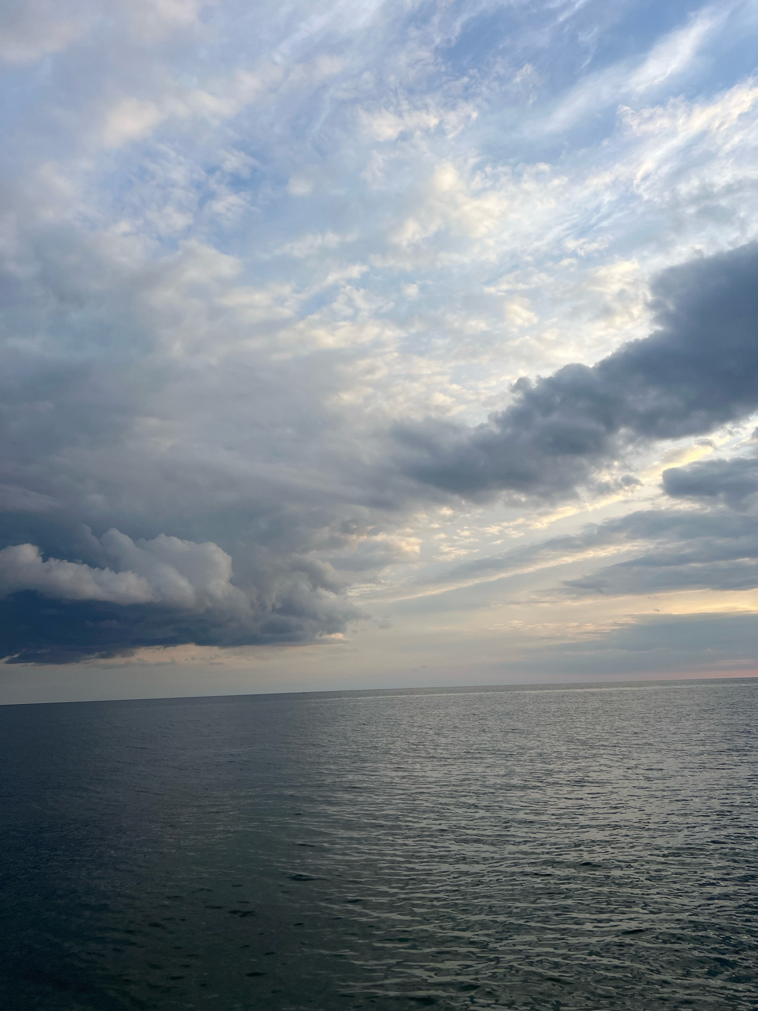 The image depicts a vast expanse of dark, rippling water meeting a hazy horizon under a dramatic sky. The sky is a mix of light and dark clouds. On the left, heavy, ominous-looking storm clouds are prominent, tinged with dark grays and purples. To the right, the clouds are lighter, with streaks of white and pale yellow where sunlight attempts to break through. The overall scene suggests a moody or transitional weather condition over the water. There are no visible people, structures, or specific landmarks to indicate a city location.