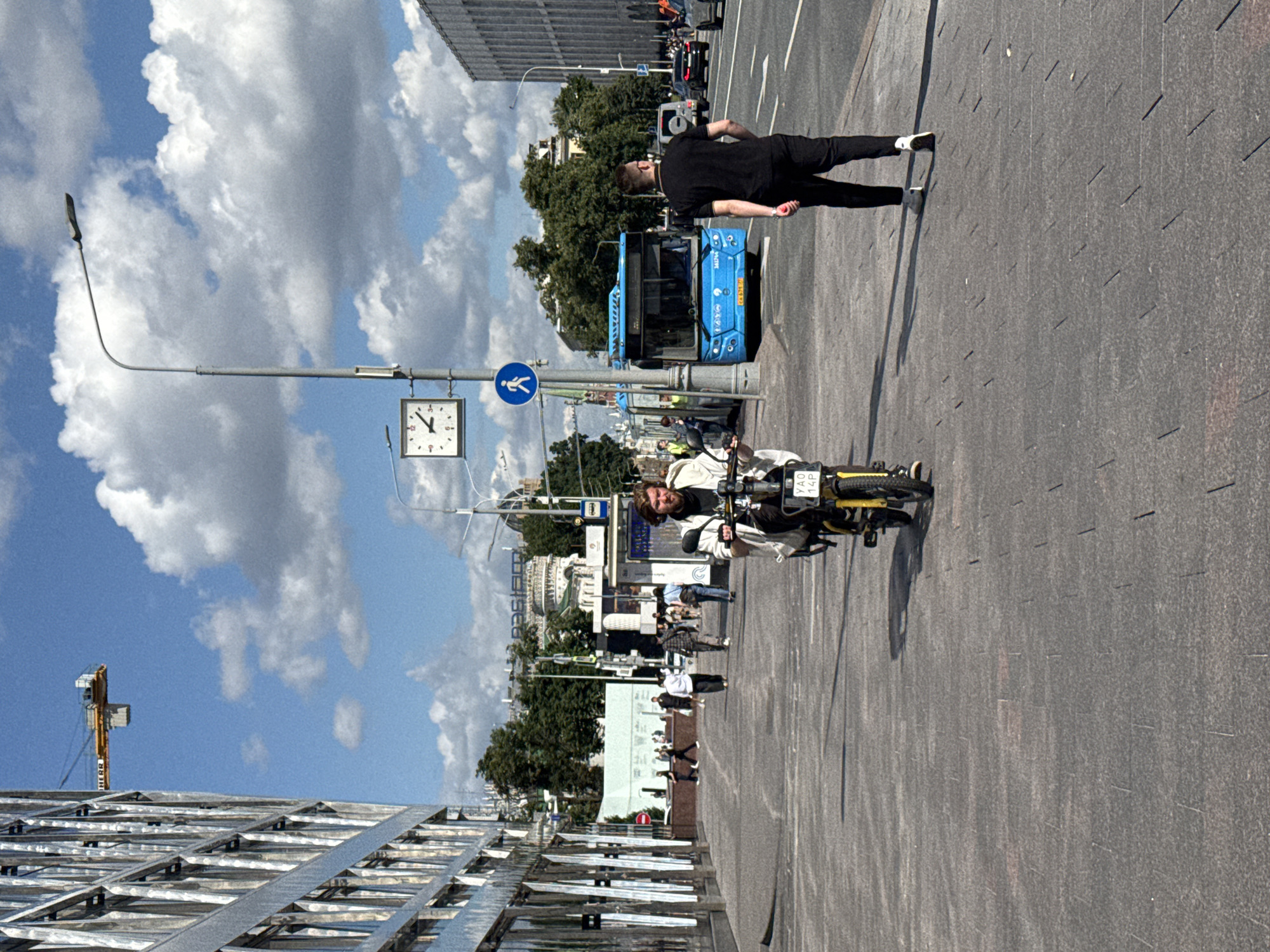 The image captures a street scene in Moscow, Russia, under a partly cloudy sky. A man is riding a yellow electric scooter on a paved pedestrian walkway. He is wearing a light-colored jacket and dark pants. To his right, a blue bus is stopped at a bus stop, with passengers boarding. A man in a black t-shirt and black pants is walking away from the viewer on the same walkway, heading towards the bus. In the background, a large modern building with reflective windows stands on the left, and another large, gray building is visible on the right. A clock is mounted on a lamppost, and a pedestrian crossing sign is visible. There are other pedestrians in the middle ground, walking towards the bus stop and along the walkway. A construction crane is visible in the sky above the building on the left. The scene appears to be a busy urban area with both pedestrian and vehicular traffic.