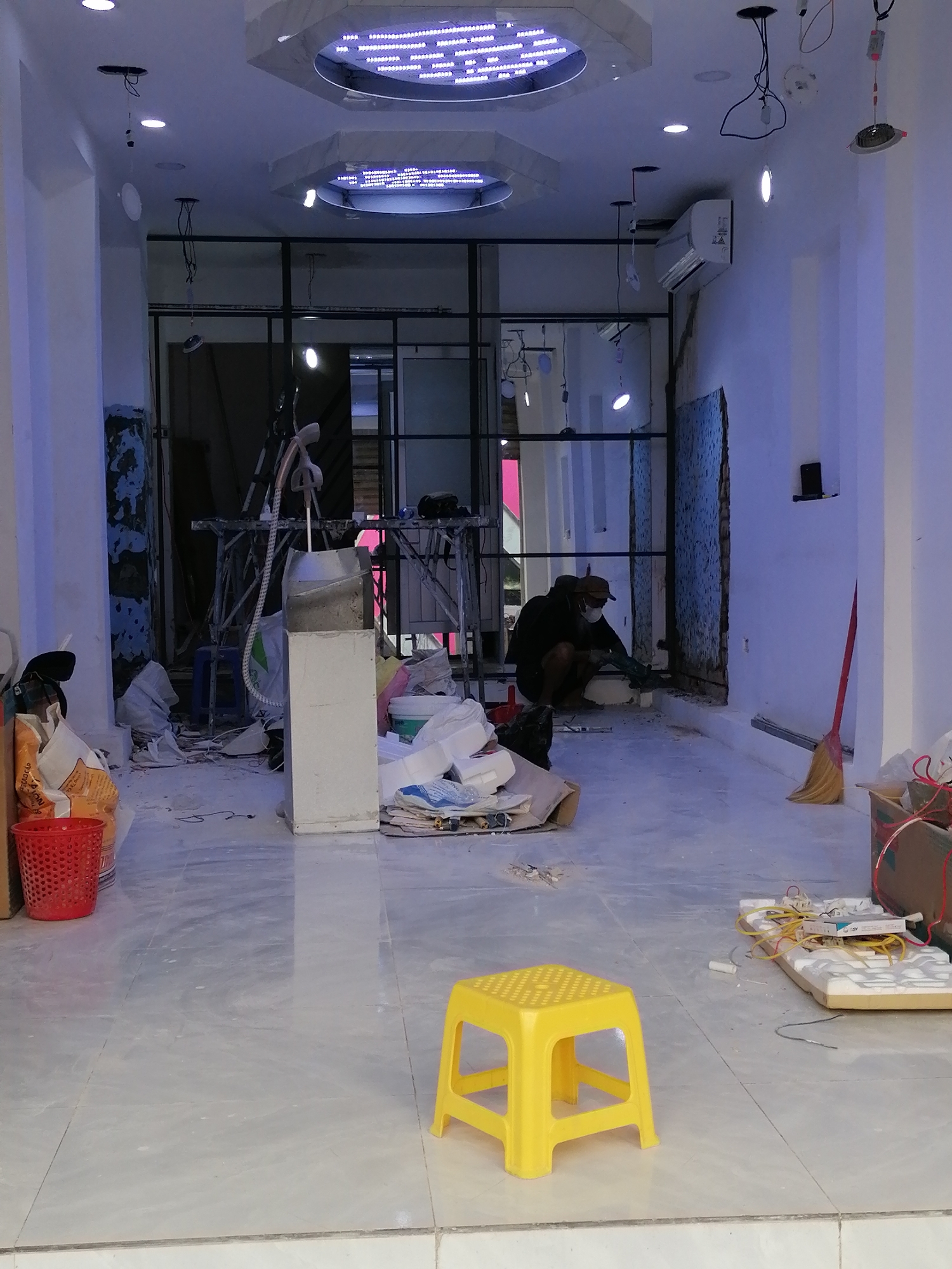 The image captures the interior of a commercial space undergoing renovations in Nha Trang Ward, Vietnam. The room is brightly lit with overhead lighting fixtures, including a distinctive circular light with LED strips. Construction materials and tools are scattered throughout the space, indicating ongoing work. A man wearing a mask is crouched near the back of the room, seemingly working on the wall. Various objects are present: a yellow plastic stool in the foreground, a red plastic bucket, a white appliance (possibly an air conditioner), construction debris, and a broom. The overall impression is that the space is being actively renovated. The storefront features glass partitions.