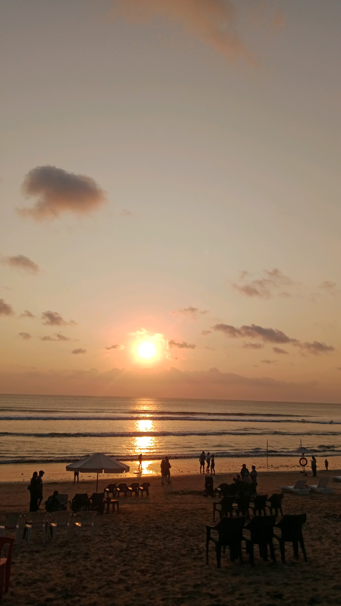 The image depicts a serene sunset scene on the beach in Kuta, Indonesia. The sun, a bright orb of light, is low in the sky, casting a warm, golden glow across the water and sand. Its reflection shimmers on the gentle waves that roll towards the shore. Scattered clouds, tinged with the colors of the sunset, dot the sky.
On the beach, several individuals are visible, some walking along the water's edge, others gathered in small groups. The silhouettes of people suggest a relaxed atmosphere. Beach furniture, including chairs and an umbrella, are present on the sand, indicating a place where visitors can unwind and enjoy the view. The sand itself is a warm, sandy hue, characteristic of a tropical beach. The overall impression is one of tranquility and the natural beauty of a sunset in Kuta.