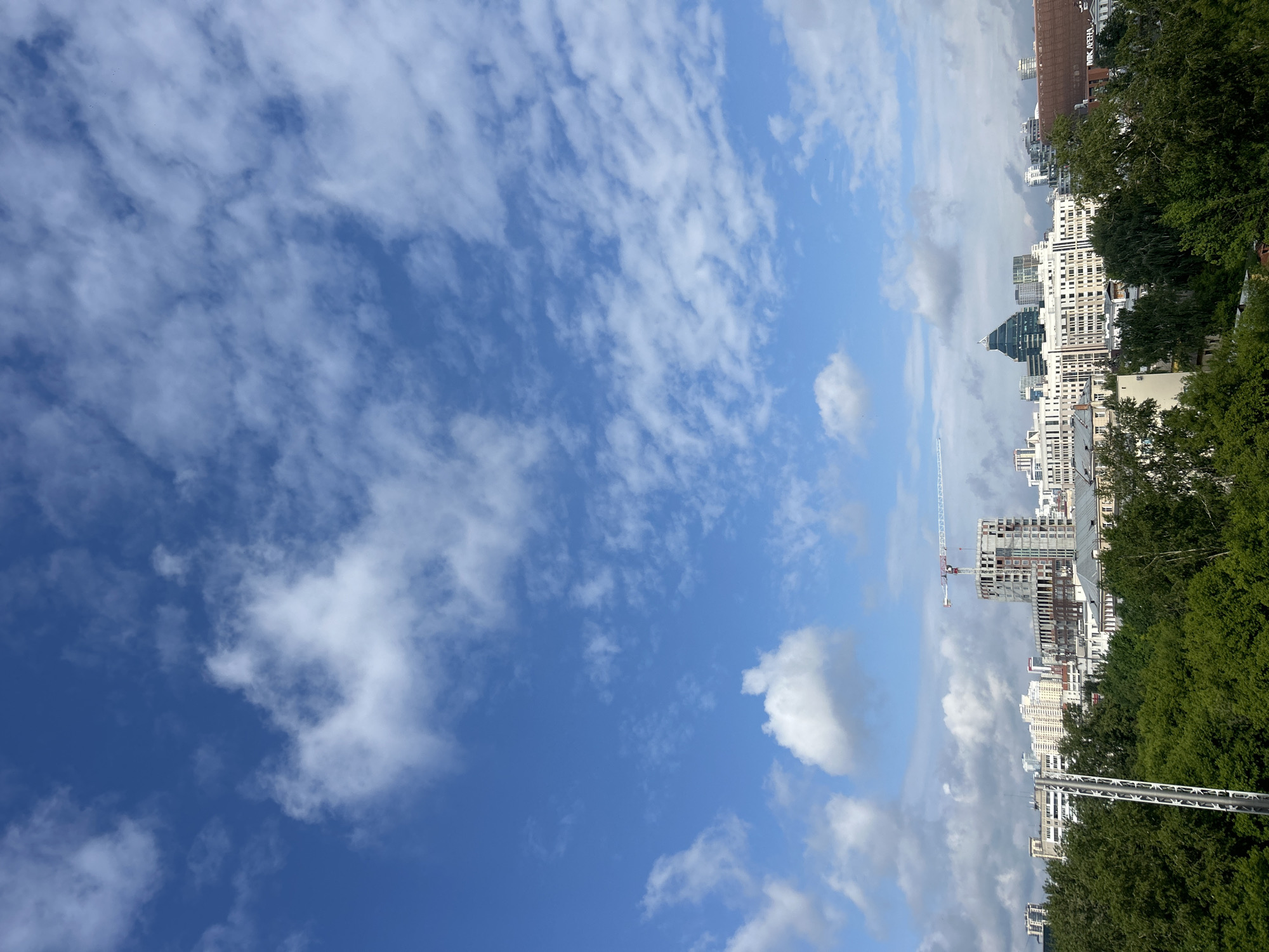 The image presents a detailed daytime cityscape view of Yekaterinburg, Russia, captured from an elevated perspective. The scene is dominated by a clear blue sky, occupying approximately the upper two-thirds of the frame, with scattered white cumulus and stratocumulus clouds of various sizes.
In the lower third of the image, the foreground is densely covered by the lush green canopy of numerous deciduous trees, indicating a park or a heavily treed urban area. On the far left, a tall, slender metallic pole structure, possibly a communication mast or light pole, extends vertically from the bottom edge into the midground.
The midground and background showcase the urban development of Yekaterinburg. Towards the center-left, a multi-story building is visibly under construction, characterized by its concrete framework and exposed rebar. A prominent red and white tower crane stands over this construction site. Adjacent and behind the trees, numerous completed multi-story buildings of varying architectural styles and colors (predominantly white, grey, and light brown) form the urban fabric.
Further to the right in the midground, a distinctive modern structure with a dark, angular, faceted roof, identifiable as the Demidov Business Center (Demidov Plaza), stands out. To its right, a large reddish-brown building with a flat roof is visible, and the Cyrillic text "АРЕНА" (Arena) is discernible on its side, indicating the Uralets Cultural and Entertainment Complex (Uralets Arena). The cityscape extends into the background with additional buildings.
No individuals or active interactions are visible in the distant scene. The overall lighting suggests bright natural daylight, consistent with morning or afternoon.