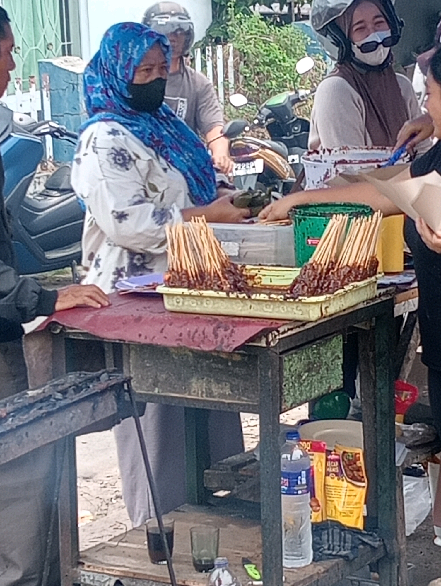 This image depicts an outdoor scene, likely a street food stall, in Bandar Lampung, Indonesia. A woman wearing a blue headscarf and a black face mask stands behind a food cart. She is in the process of preparing skewers of meat, which are neatly arranged in a rectangular tray. To her left, another person, partially visible, is handling what appears to be food preparation. Behind them, a man wearing a helmet is seated on a motorcycle. The food cart itself is a mobile unit with a metal frame, holding various items including a green bucket, plastic containers, and packaged food products. Several glasses of liquid are also visible on the cart. The background suggests a moderately populated area, with glimpses of buildings and greenery. The lighting indicates it is daytime.