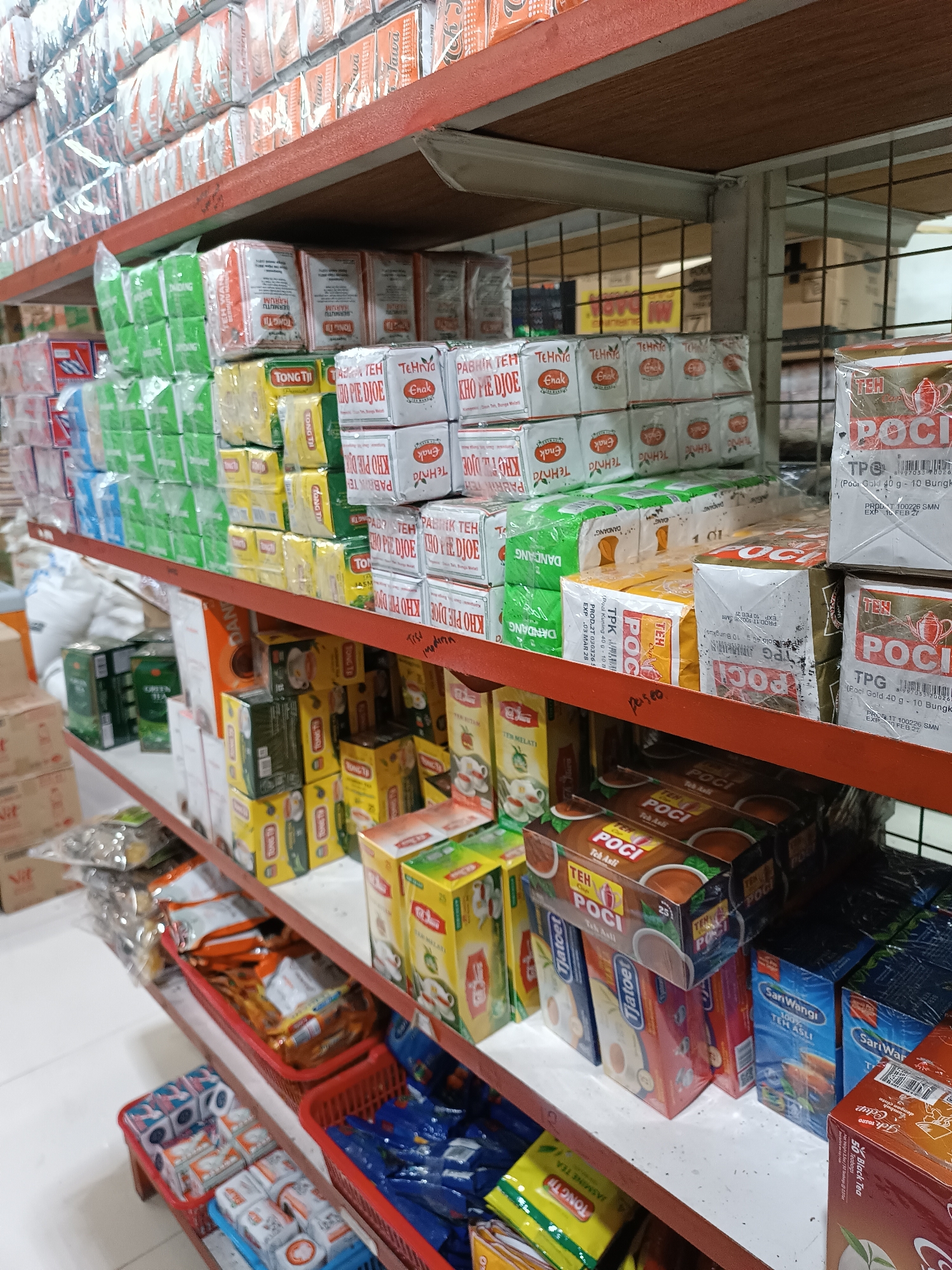 This is a full, eye-level shot of a retail store aisle in Jetis, Indonesia, specifically a section of shelves stocked with various tea products. The shelves are made of wood and metal, and the lighting is bright and even, suggesting an indoor commercial environment. The products are predominantly tea boxes and packets, with prominent brands like "POCI," "TONG TI," "Dandang," and "SariWangi" visible. Some boxes are stacked neatly, while others are arranged in red plastic baskets. There are no people visible in the image. The aisle appears to be a typical grocery or convenience store setting, with an organized but densely packed display of merchandise.