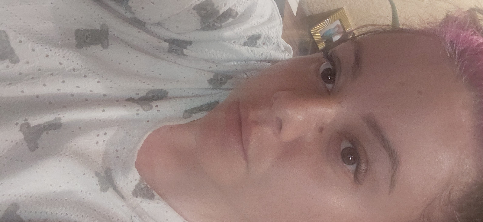 The image shows a close-up, selfie-style photograph of a woman's face. She has light skin and is wearing a white top with a gray teddy bear pattern. Her hair is styled up, with some pink-colored strands visible at the top of her head. She has dark eyebrows and is wearing what appear to be false eyelashes. The background is blurry and appears to be an indoor setting, possibly a home. A small framed picture is visible on the left side of the image, in the background. The lighting suggests it is daytime, but the source of light is not clear. The photograph was taken in Marynivka, Ukraine.