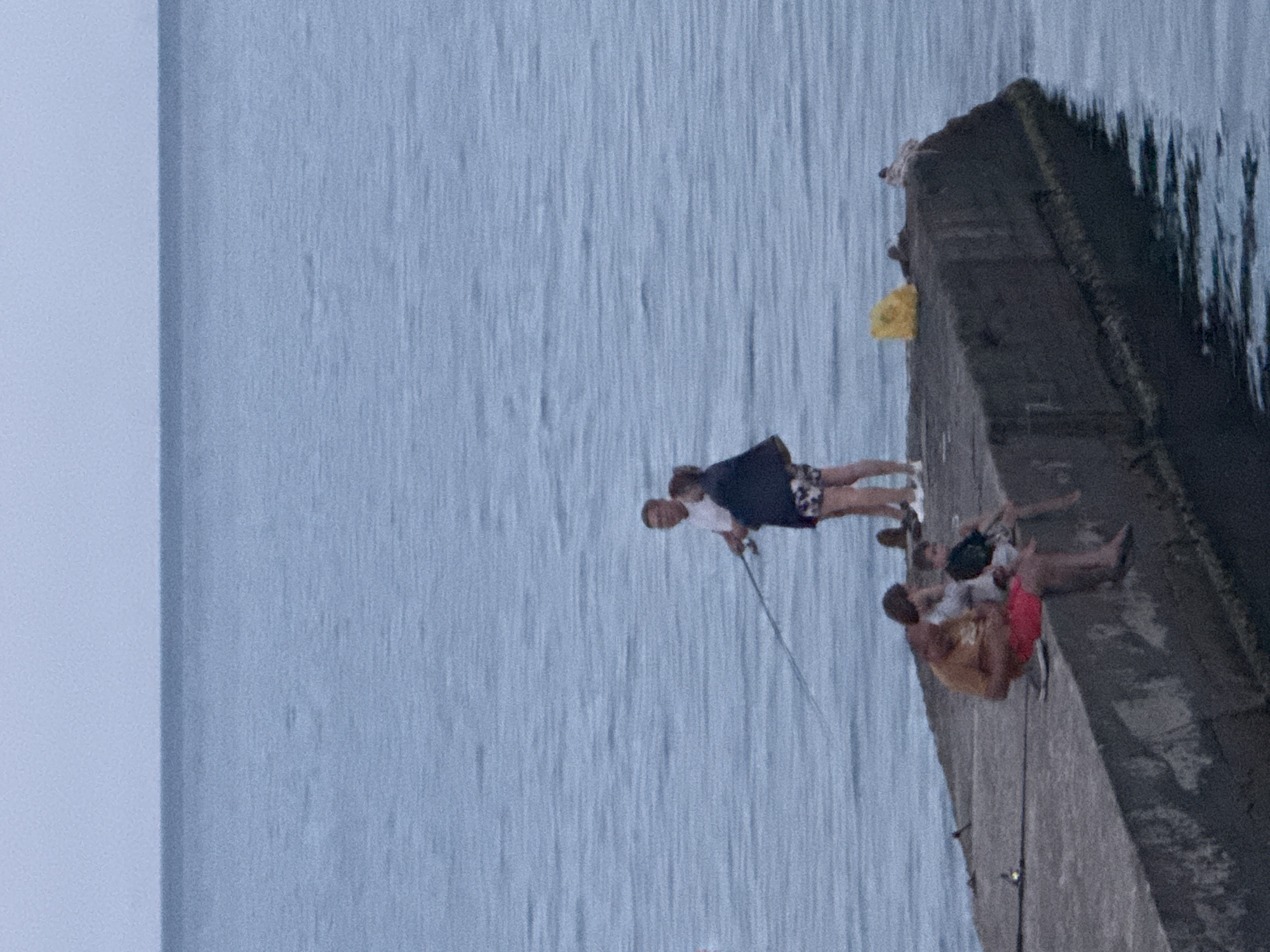 The image shows a concrete pier extending into a calm, blue body of water, likely the Black Sea, near Partenit, Ukraine. The sky is overcast and light blue. On the pier, two adults and two children are visible. Towards the back, a man in a white shirt and patterned shorts stands with a fishing rod, and a woman in a dark jacket and patterned shorts stands beside him. In the foreground, a man in a yellow shirt and red shorts sits with his legs extended, and a child in a dark t-shirt sits beside him, with their legs dangling over the edge of the pier. Another child is partially visible between them. The pier is weathered and shows signs of age. A yellow plastic bag and a bird are on the pier near the edge.