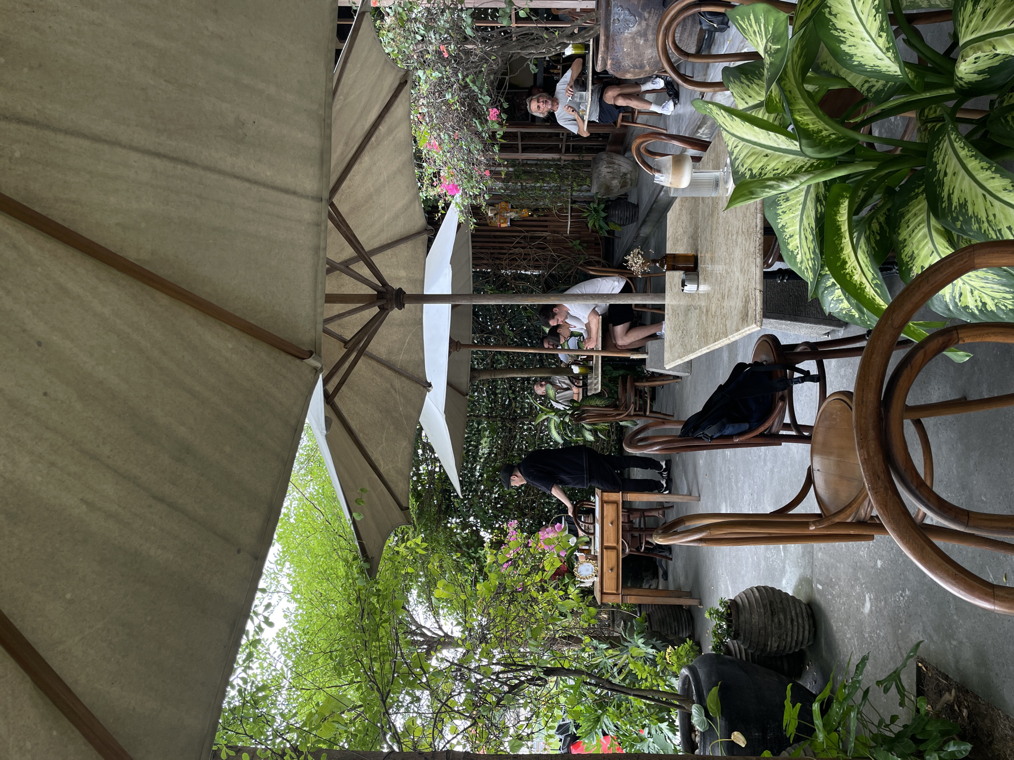 The image depicts an outdoor seating area of a cafe in North Kuta, Indonesia. Several people are seated at tables, some are dining or conversing, while others appear to be relaxing. A staff member in a black outfit is standing near a wooden desk. The area is adorned with lush greenery, potted plants, and outdoor umbrellas providing shade. The overall atmosphere suggests a casual and relaxed dining experience.