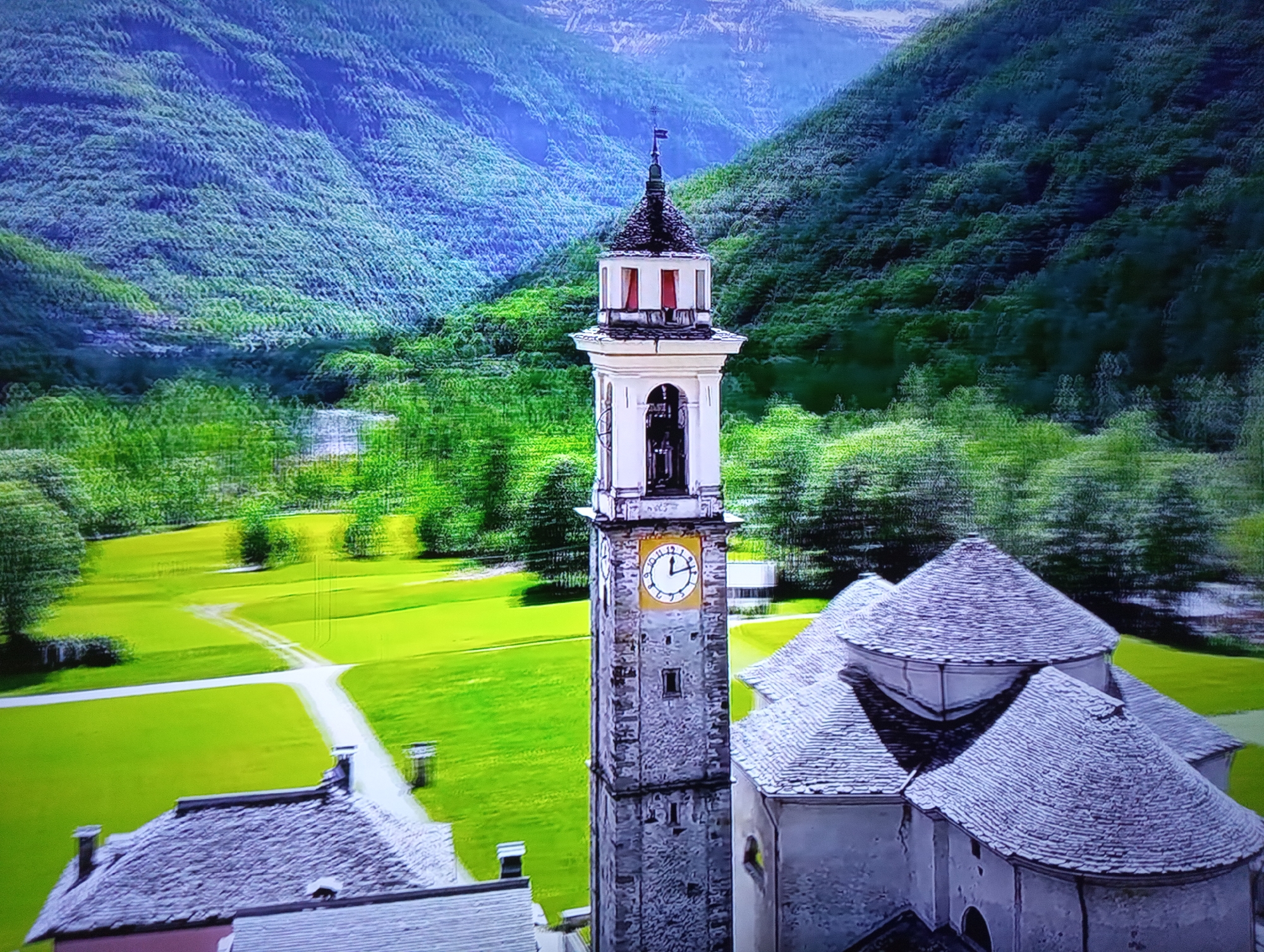 The image depicts an aerial view of a stone clock tower and an adjacent building with multiple conical roofs, set against a backdrop of lush green, terraced hills and mountains. The clock tower has a white facade with red-trimmed windows and a clock face that shows the time as approximately 1:50. In the foreground, parts of other stone-tiled roofs are visible. A winding path or road leads through a vibrant green field. The scene suggests a rural or mountainous location, possibly a historical or religious site. There is no indication of the city of Dnipro, Ukraine, within the image.