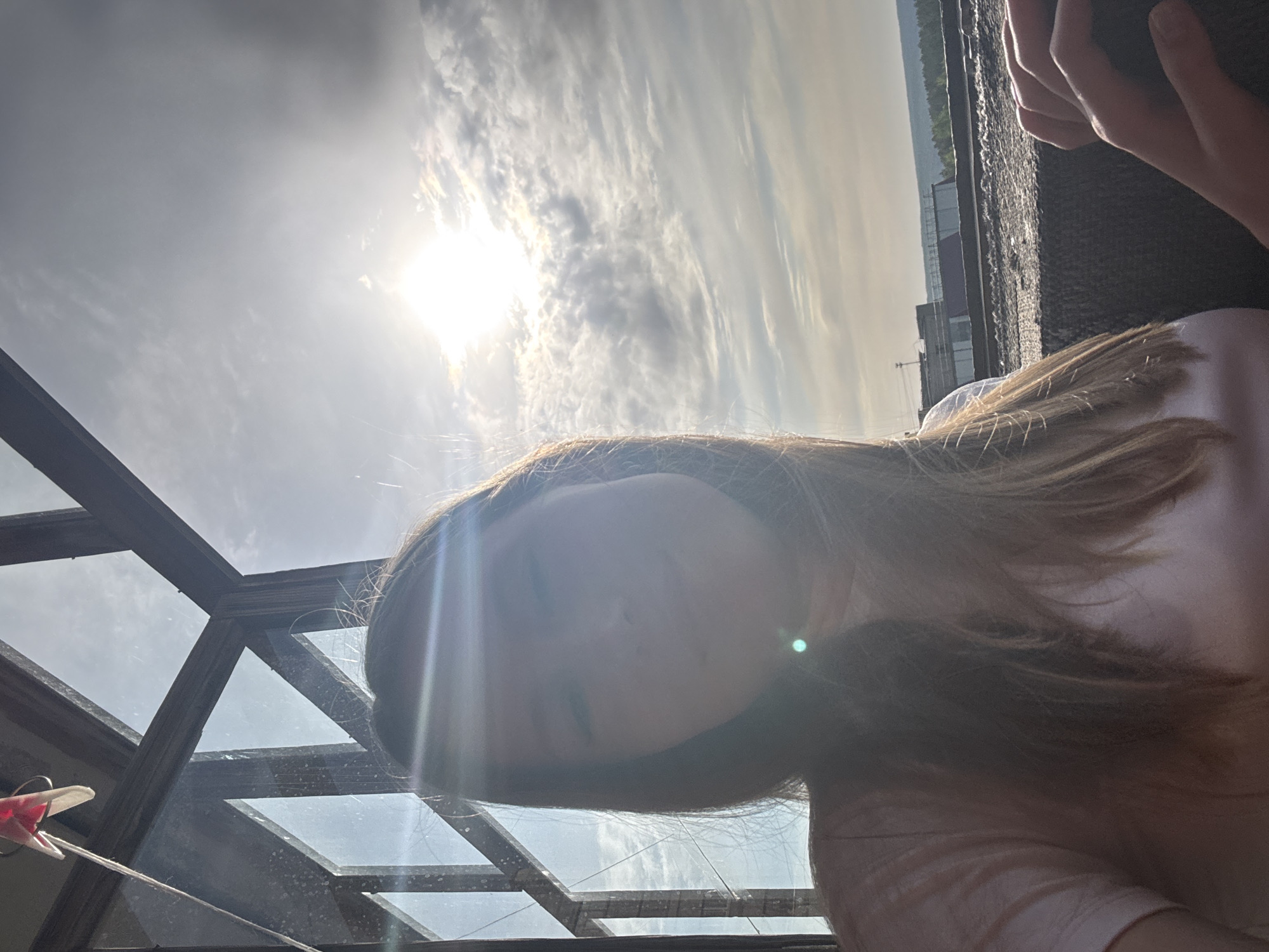 The image presents a view of a person with long brown hair, partially obscuring their face due to the bright sunlight. The individual appears to be indoors, looking up through a glass roof structure with a wooden frame. Above, a sky with bright sun and clouds is visible. Through the glass, outlines of buildings and trees are visible in the distance. A hand is visible in the upper right corner, seemingly holding a phone or camera. To the left of the image, a small airplane model hangs from a string. Without specific landmarks or contextual clues, a definitive location within Chaikovsky, Russia is not possible.