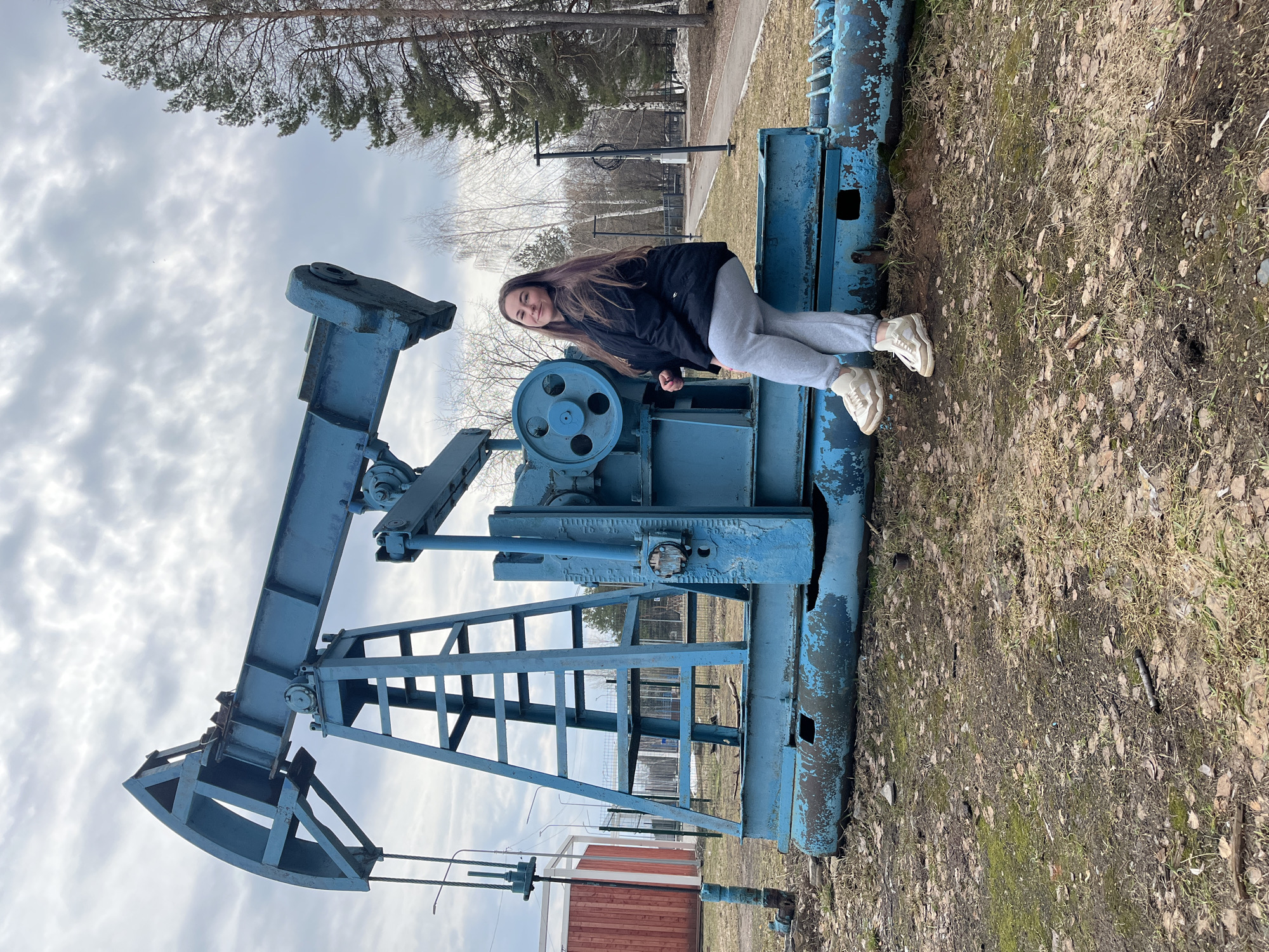 A young woman is posing on an old, blue pumpjack oil well in Chaikovsky, Russia. She is wearing a black jacket, gray sweatpants, and light-colored sneakers. The pumpjack is located in an outdoor area with trees and a cloudy sky. The ground is covered with dry grass and leaves. A small, red building is visible in the background.