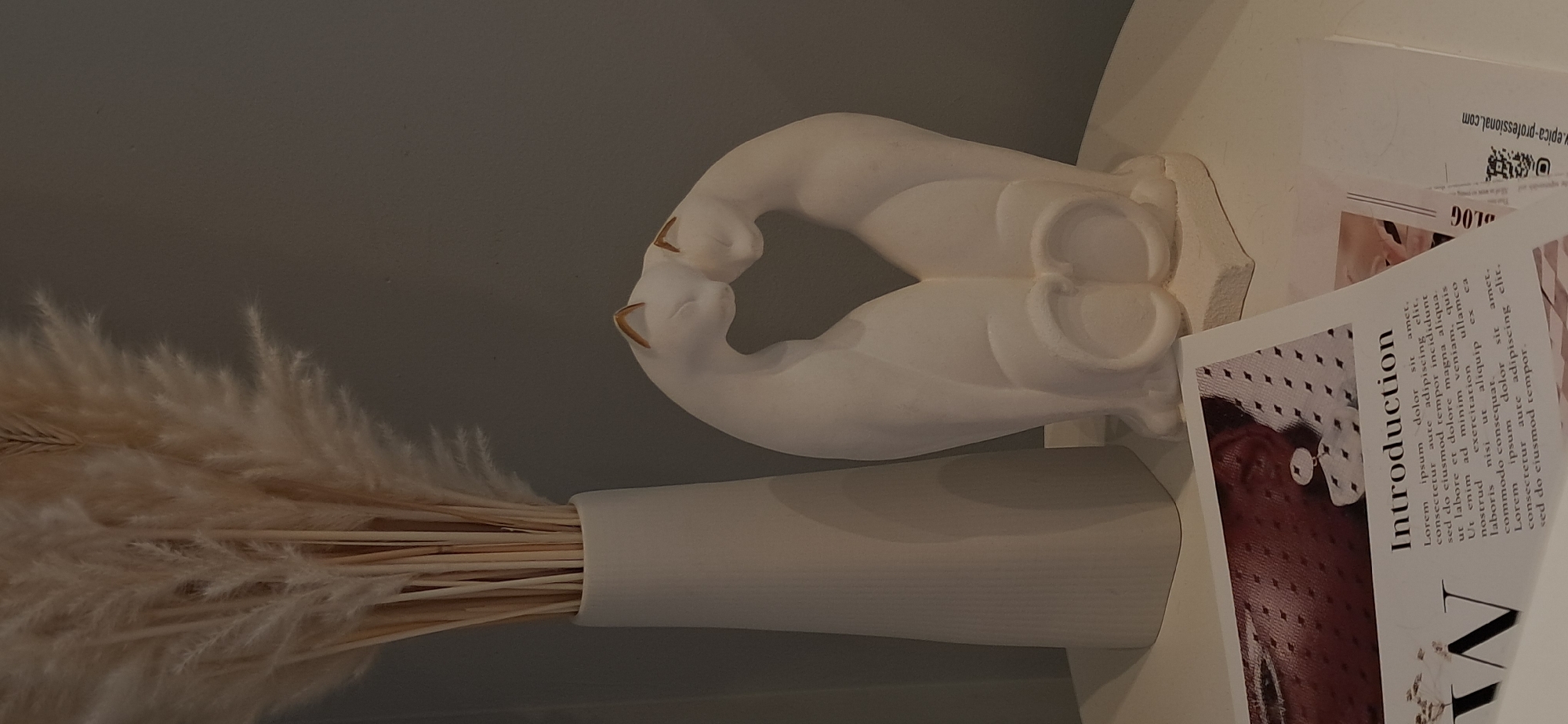 A still life scene features a white vase with dried reeds or pampas grass, a white sculpture of two cats embracing, and a magazine on a white surface, against a muted gray wall. The magazine, angled to the right, has the title "Introduction" printed in black text above columns of Latin placeholder text. A portion of a person's face, wearing an earring, is visible on the magazine's left page. A second, partially obscured document is to the right of the magazine, showing text that includes "LOG" and a web address "epica-professional.com". The scene is lit by an overhead light source, casting soft shadows. The overall aesthetic is minimalist and serene. This image was taken in Moskva, Russia.