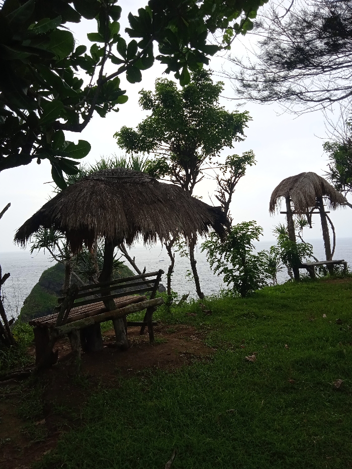 The image shows a scene of a grassy cliff overlooking the ocean in Girisubo, Indonesia, with a thatched roof hut and a wooden bench in the foreground. The hut is made of woven palm leaves and has a thatched roof. The bench is made of wooden planks and has a natural wood finish. The hut and bench are situated in the shade of a large tree. Behind the bench is a small rocky outcrop and a grassy area. There are two smaller trees in the background, both with a similar thatched roof design. The ocean can be seen in the distance, and a small boat is visible in the water. The sky is overcast and grey. The scene is quiet and peaceful.