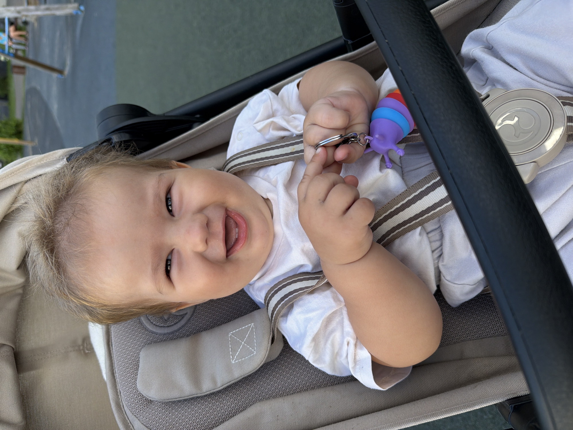 A close-up, eye-level shot captures a smiling baby, likely under one year old, seated in a stroller. The baby has light brown hair and is looking towards the camera with bright eyes and an open mouth, showing teeth. They are wearing a white t-shirt with brown and white striped suspenders and light gray pants. The baby's left hand is near their mouth, holding a keychain with a colorful caterpillar-shaped toy. The stroller is beige with gray padding and a black safety bar in the foreground. In the blurred background, a playground with dark gray rubber surfacing and wooden play structures is visible, suggesting an outdoor setting. The overall lighting is bright, indicating daytime. The location is specified as Kazan, Russia.