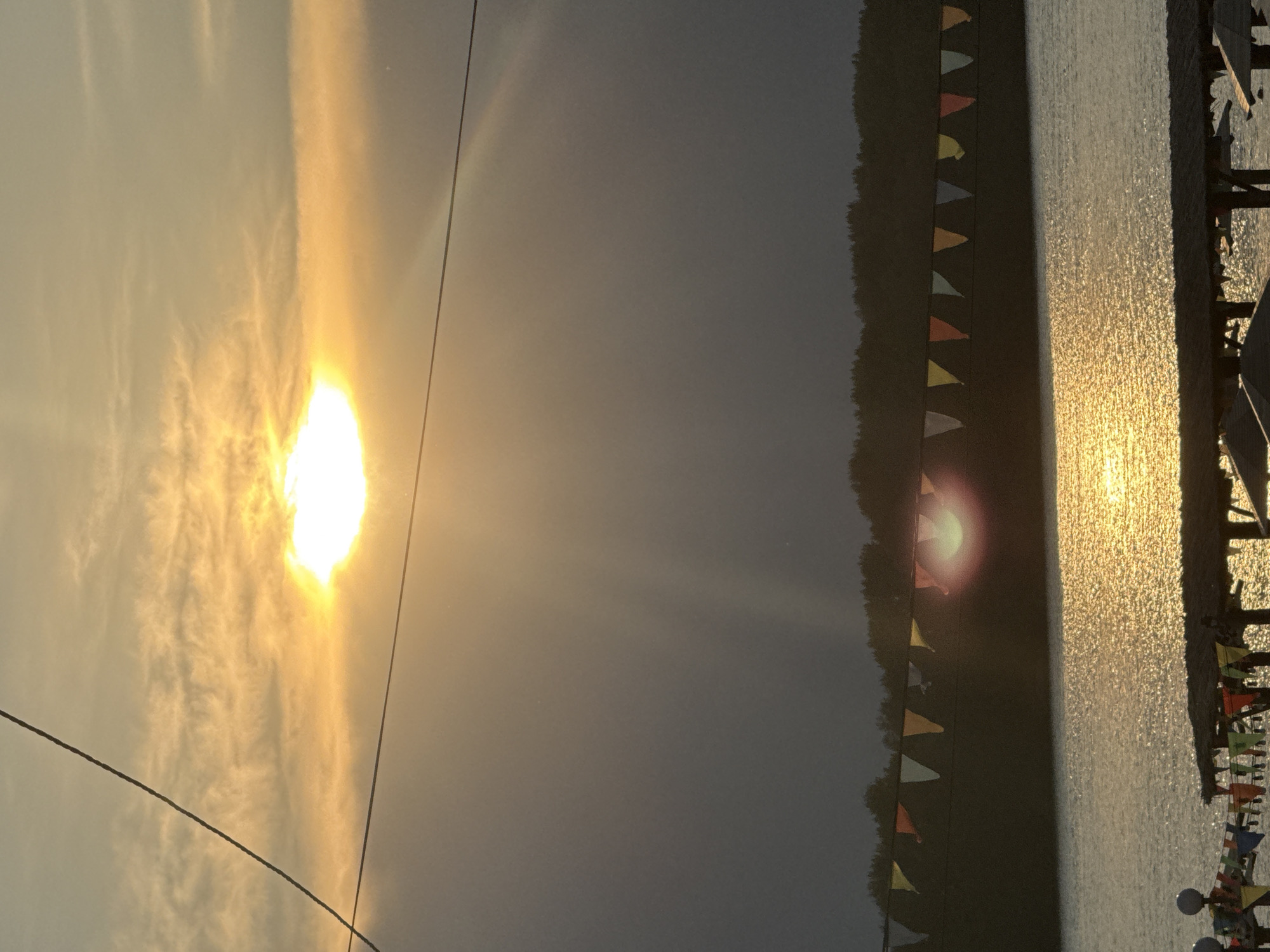 The image shows a body of water, possibly a river or lake, reflecting the golden light of a sunset. A dark tree line borders the far side of the water. Above the water, a string of colorful triangular flags is visible, strung horizontally. In the sky, the sun is setting behind clouds, casting rays of light. The overall scene suggests an outdoor recreational area, perhaps a beach or waterfront park. The specific location in the city of Pavlodar, Kazakhstan, cannot be determined from the image alone.