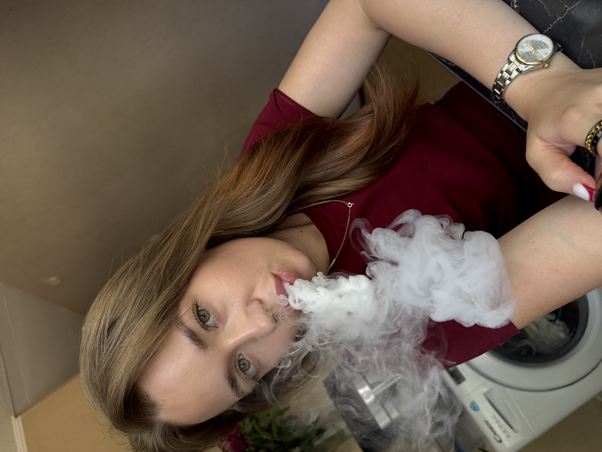 A woman with long, light brown hair and green eyes is exhaling a cloud of white vapor. She is wearing a dark red top, a gold and silver watch on her left wrist, and a ring on her right hand. The photo appears to be taken indoors, possibly in a laundry room, as a washing machine is visible in the background. The lighting suggests it could be daytime, but the exact time is not discernible. The woman's expression is neutral. No discernible text is visible in the image. The orientation of the image is rotated.