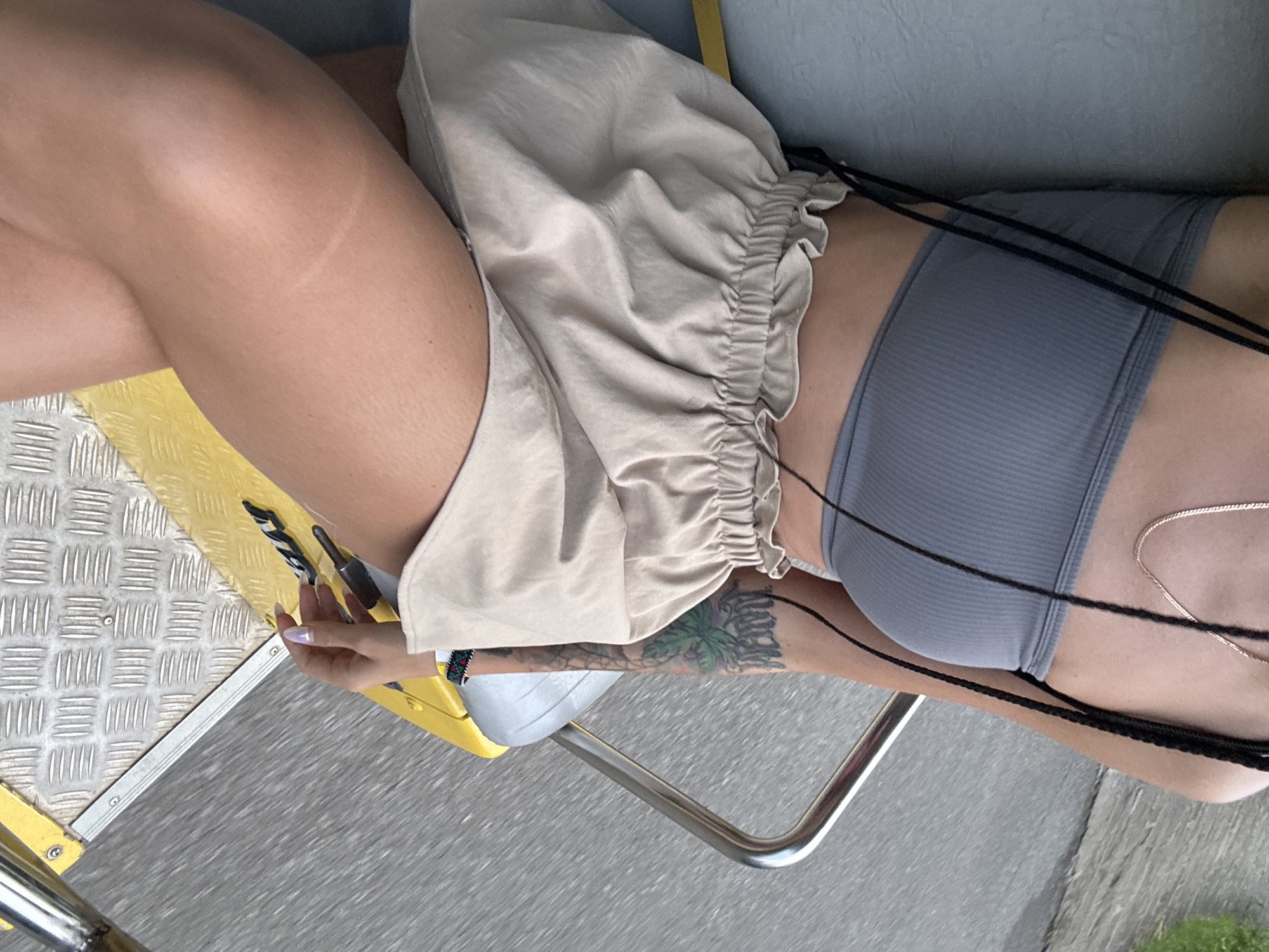 The image shows a close-up of a young woman sitting on a yellow bench outdoors. She is wearing a grey strapless top and light-colored ruffled shorts. Her left arm, visible in the frame, has a tattoo of a palm tree and some text. She is also wearing a gold chain necklace and has dark braids. The background appears to be a blurry outdoor scene with gray asphalt. The woman is seated on what looks like a public transportation vehicle or park seating. The specific location of Tây Hoa Lư Ward, Vietnam is not visually identifiable in the image.