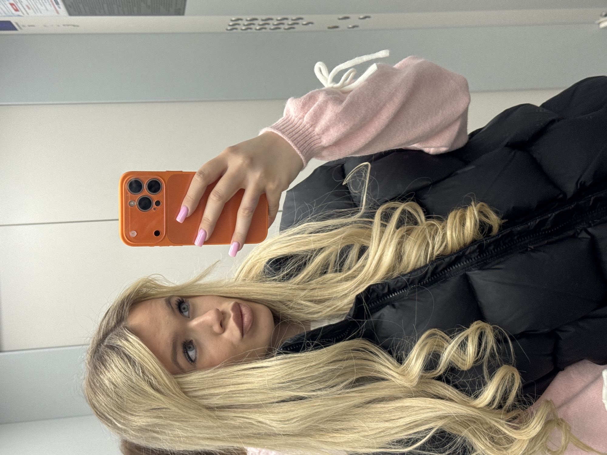 A young woman with long blonde wavy hair is taking a mirror selfie. She is wearing a black puffer vest over a light pink sweater with white tie details on the sleeves. She has light blue eyes and is wearing light makeup. She is holding an orange smartphone in her right hand. The background appears to be an indoor setting, possibly a hallway or elevator, with light gray walls. There are some notices or advertisements visible on a vertical surface to the right of the frame, suggesting a public or commercial space. The overall lighting is neutral, and the image is taken from the chest up. The location is stated to be Sapernyy, Russia.