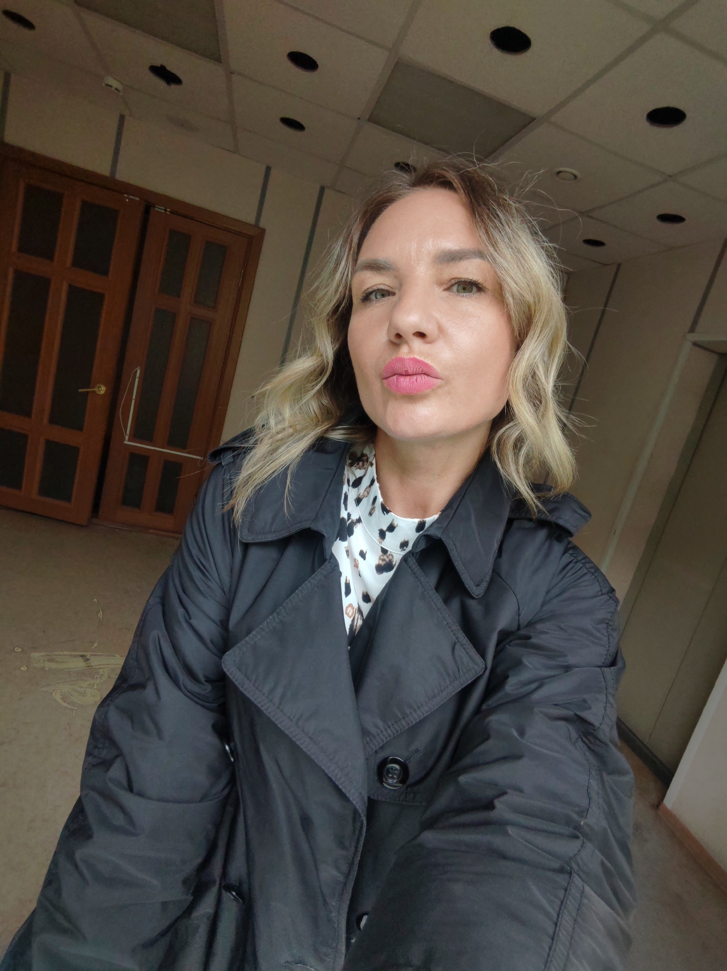 A woman is taking a selfie indoors. She has blonde hair with dark roots, green eyes, and pink lipstick. She is wearing a black trench coat over a patterned shirt. Behind her are closed brown wooden doors and a drop ceiling with recessed lighting. The location is likely indoors, but not identifiable to a specific location in Petropavlovsk-Kamchatsky.