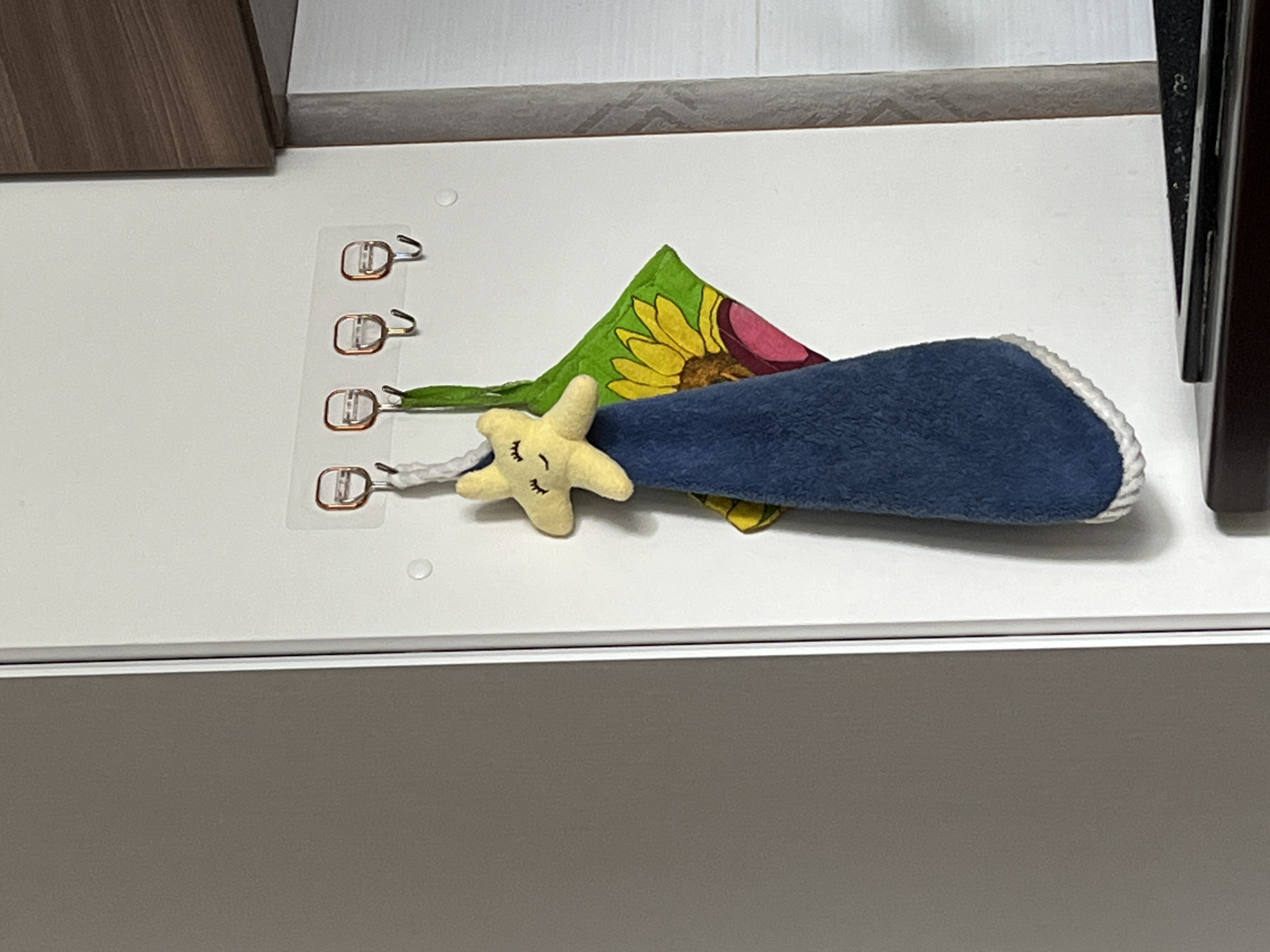 A plush yellow star with embroidered eyes and a smiling mouth hangs from a white string. The string is attached to a clear adhesive hook, one of four on a clear plastic strip adhered to a white wall. A blue towel with a white rope-like trim hangs from the star. To the right of the star, a green cloth with a painted yellow sunflower and pink petals is also hanging from a hook. The scene is lit by artificial light. The background shows a grey panel on the left, a white wall with wooden trim at the top right, and a dark surface at the bottom.