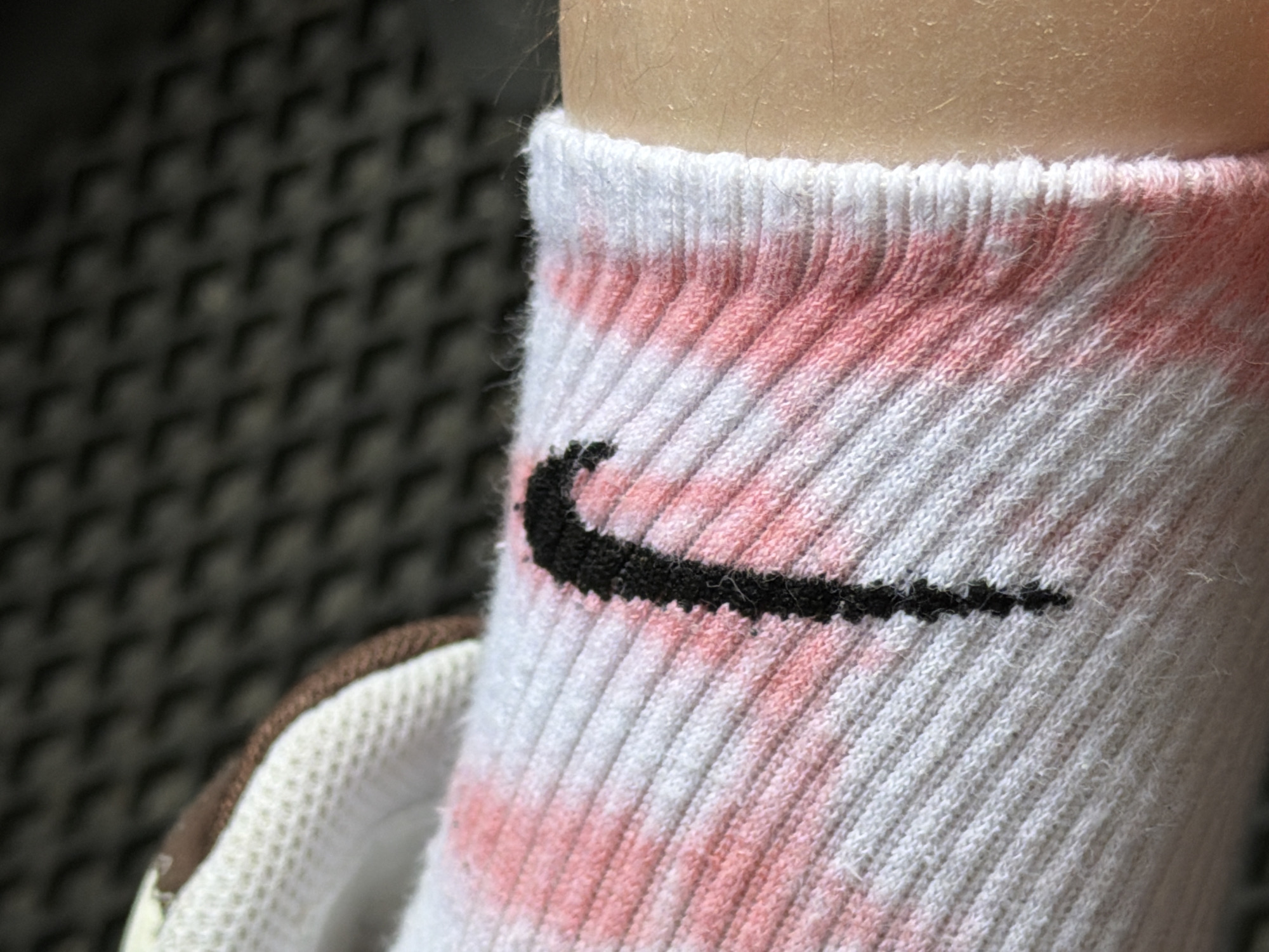 The image shows a close-up of a person's leg wearing a white sock with a black Nike logo. The sock has pinkish streaks of color, possibly from dye transfer or a stain. The background is out of focus and appears to be a textured surface, possibly outdoor flooring. The location in the city of Катунь, Russia, is not discernible from the image itself.