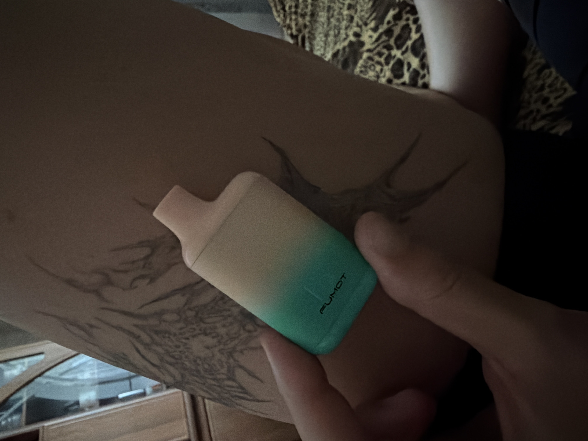 A close-up, low-angle shot shows a person's hand holding a small, gradient-colored vape device. The vape has the word "FUMOT" printed on its side in black lettering. The person's arm is visible in the background, adorned with a large, dark tattoo of abstract, flame-like shapes. The individual is lying down on a surface covered with a leopard-print fabric, and a piece of dark fabric is visible in the lower portion of the frame. The environment appears to be indoors, with a hint of furniture and possibly a window in the upper left background.