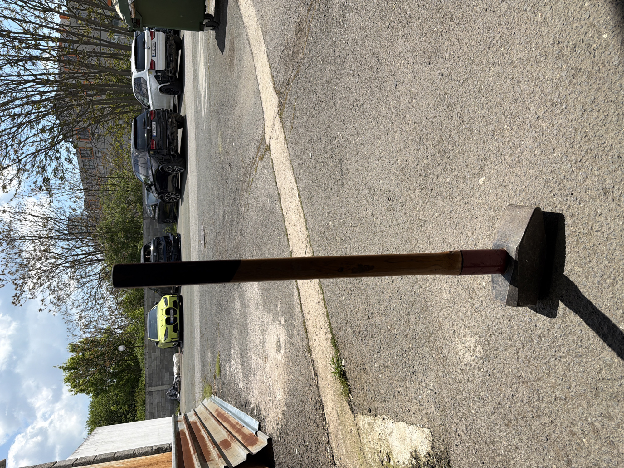 A sledgehammer lies on asphalt pavement in an outdoor urban environment. The sledgehammer has a dark metal head and a long brown wooden handle with a burgundy accent near the head. Cars are parked in a row in the background, ranging in color from white to black to lime green. A green garbage container is also visible. The scene appears to be in a parking lot or alleyway. A building and trees are in the background. The sky is cloudy. The location is Burgas, Bulgaria.