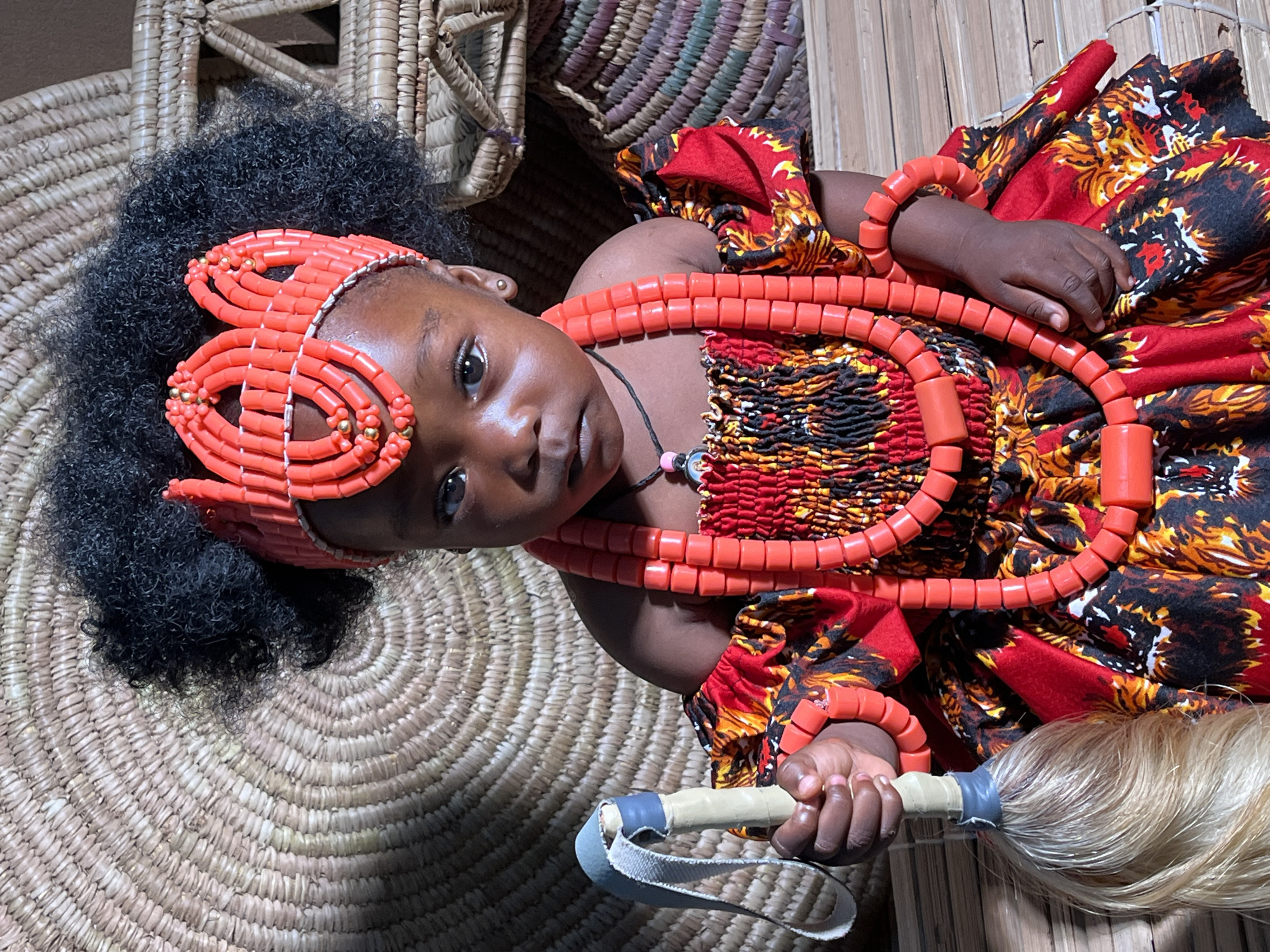 A child, likely female, is dressed in traditional Nigerian attire. She has dark hair with a coral-colored beaded headpiece. She also wears multiple strands of coral beads around her neck and on her arms. Her dress is a vibrant mix of red, yellow, and black patterns. She holds a ceremonial whisk with a white handle and light-colored fibers. The child is positioned on a woven mat, possibly a traditional Nigerian design. A woven basket is visible in the background. This is possibly located in the city of Barakin Akawo, Nigeria.