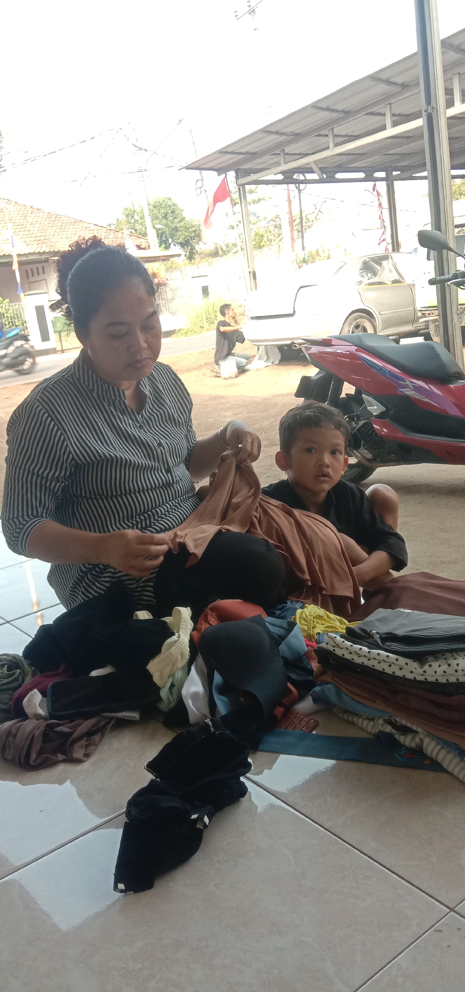 In Subang, Indonesia, a woman with her hair in a bun, wearing a black and white striped shirt, sits on the ground sorting laundry. Beside her, a young boy in a black shirt watches. A pile of clothes, including bras, is spread between them on the tiled floor. In the background, a motorcycle is parked under a metal awning, and a car is visible undergoing repairs or detailing. The scene takes place outdoors, with a house and trees visible further in the distance.