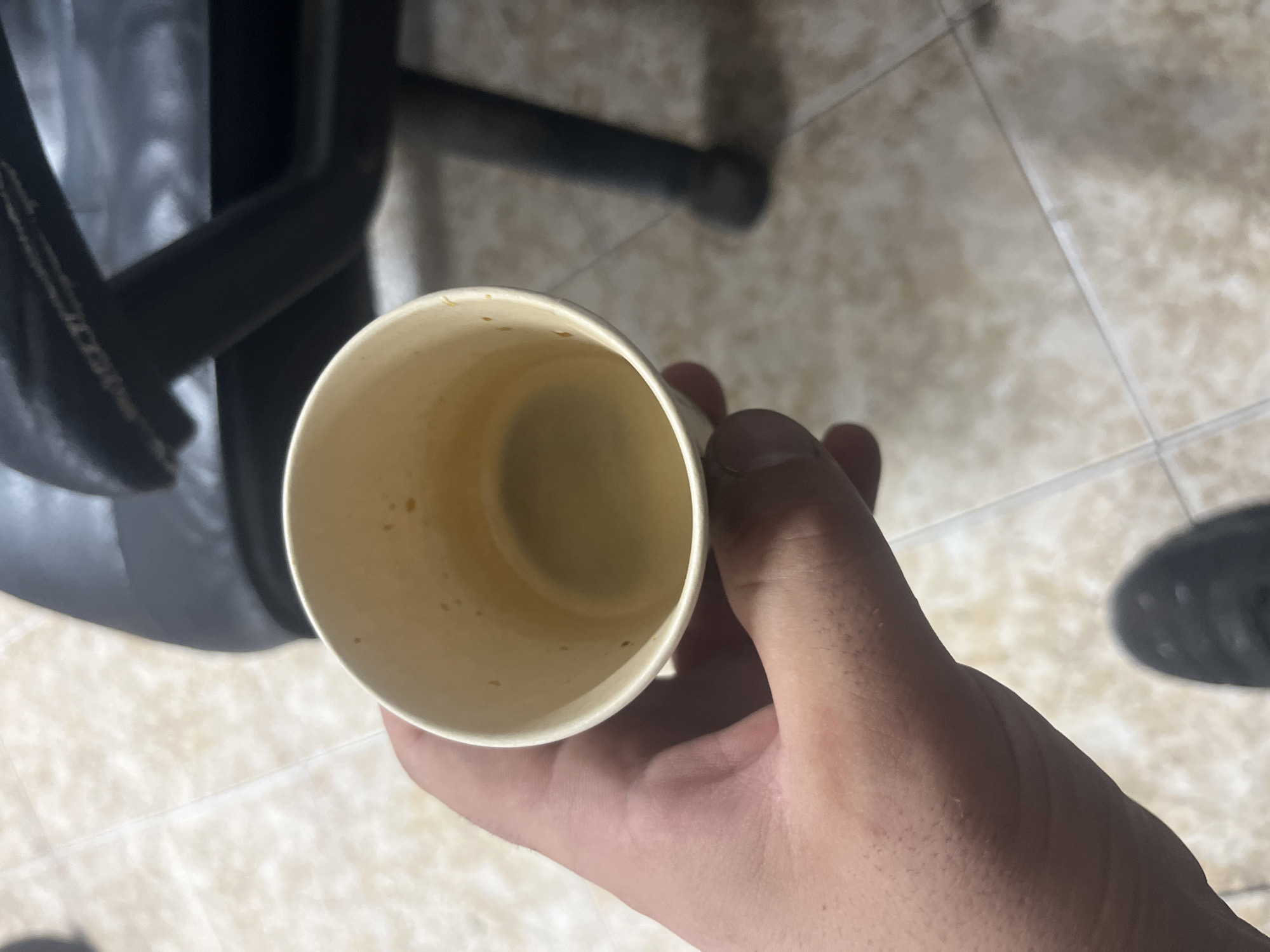 A person's hand is holding a partially full paper cup of a light brown liquid, likely coffee or tea. The cup is light-colored with some dark specks visible on the inside rim. The background shows a beige tiled floor and the dark leg of a chair. The scene is indoors.