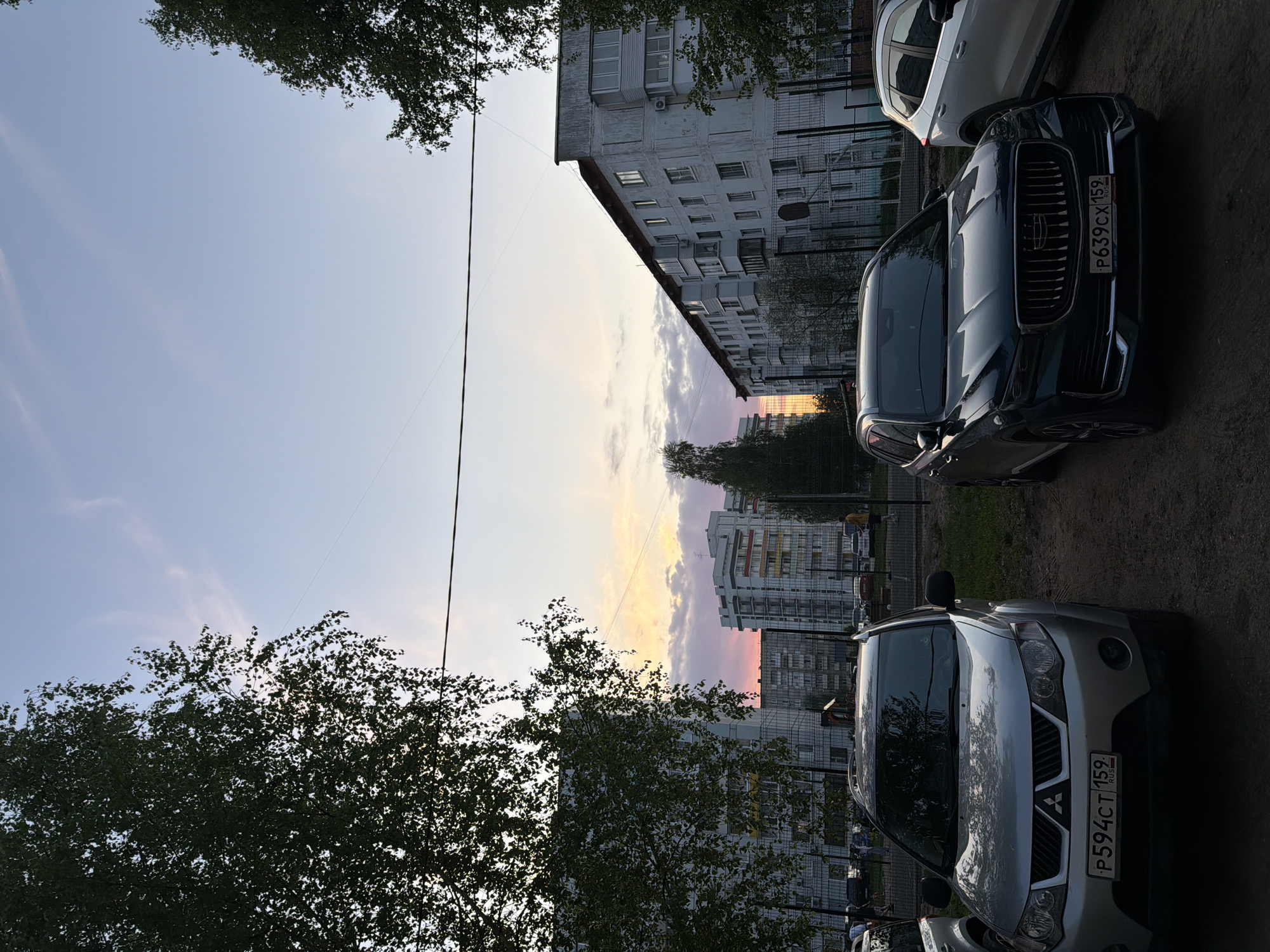 The image shows a street scene in Chaikovsky, Russia at dusk. Cars are parked on both sides of the street. A silver Mitsubishi Outlander with license plate P594CT159 and a dark-colored SUV with license plate P639CX159 are prominently visible. Buildings line the street, partly obscured by trees. Overhead, power lines run across the sky. The sky shows colors of sunset.