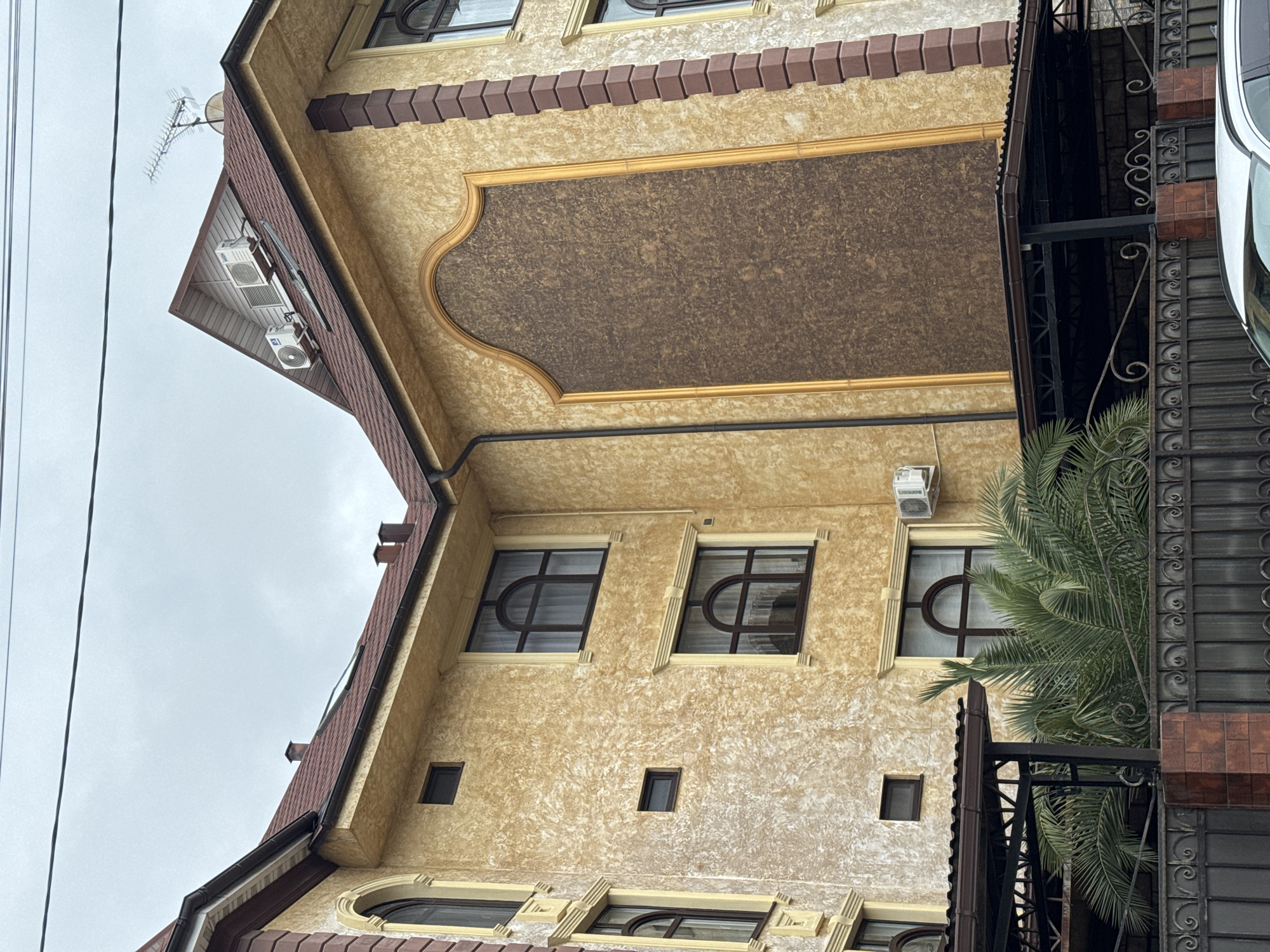 This media file captures a multi-story building, likely a residence or guesthouse, in Sochi, Russia, under an overcast sky. The building features a textured, light golden-beige stucco facade with ornate architectural details. Its roof is a dark reddish-brown, possibly tiled, and includes brown gutters and downspouts.
Various windows are visible, some rectangular and others featuring arched or semicircular tops within rectangular frames, all with dark sills and decorative surrounds. A prominent decorative element on the upper facade is a large, central rectangular panel framed in gold, with an ornate, curved top edge and a dark brown, finely textured surface. Below the roofline and above some windows, there is a repeating pattern of brown, stepped decorative blocks. Several white outdoor air conditioning units are mounted on the exterior, including two on the gable end of the roof, where a television antenna and a satellite dish are also present.
To the right, a vibrant green palm tree partially obscures the building and an ornate dark metal fence or gate with intricate scrollwork, which also incorporates brown brickwork. Part of a white car is visible behind the fence on the far right. Dark power lines stretch across the upper left portion of the grey, cloudy sky. The scene is static, depicting no explicit human activity. The time of day is daytime, characterized by the natural but diffused light of an overcast day. No visible text is present in the image.