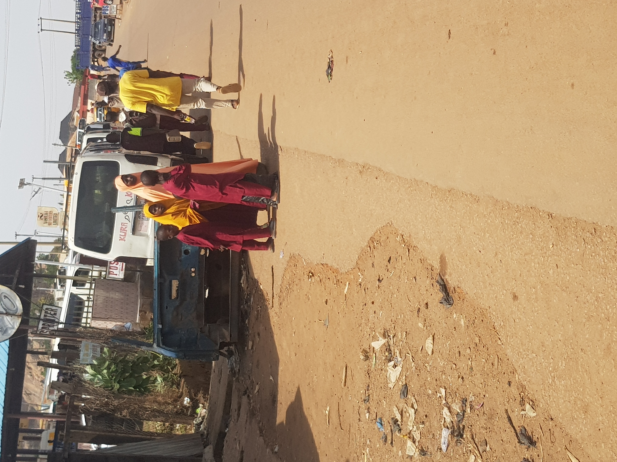 This image captures a vibrant street scene in Kufena, Nigeria, under bright sunlight, indicated by the long, distinct shadows suggesting either early morning or late afternoon. The setting is an unpaved road with sections of asphalt, flanked by buildings and sparse vegetation.
In the foreground, a group of three children or young people stands near a white van, two wearing red robes and one in an orange-yellow robe and head covering, possibly indicating religious attire or school uniforms. Next to them is a partially disassembled blue vehicle, resembling an old car chassis. Litter, including plastic bags and debris, is scattered along the dirt shoulder of the road.
Behind the robed individuals, several people are visible. A man in a bright yellow shirt and light-colored pants walks in the mid-ground. Further back, more individuals are seen, some near parked vehicles. A person in a blue shirt is in the distance. The street is active with various vehicles, including several vans and what appears to be a blue flatbed truck in the background.
Visible text includes "KURDA" and "POS" on the white van. A sign for "KASHAWA SOVENTURES LIMITED" is legible, along with another sign that reads "SPRITER". Utility poles and power lines crisscross the upper part of the scene. The overall atmosphere depicts a bustling street in an urban or semi-urban Nigerian environment.