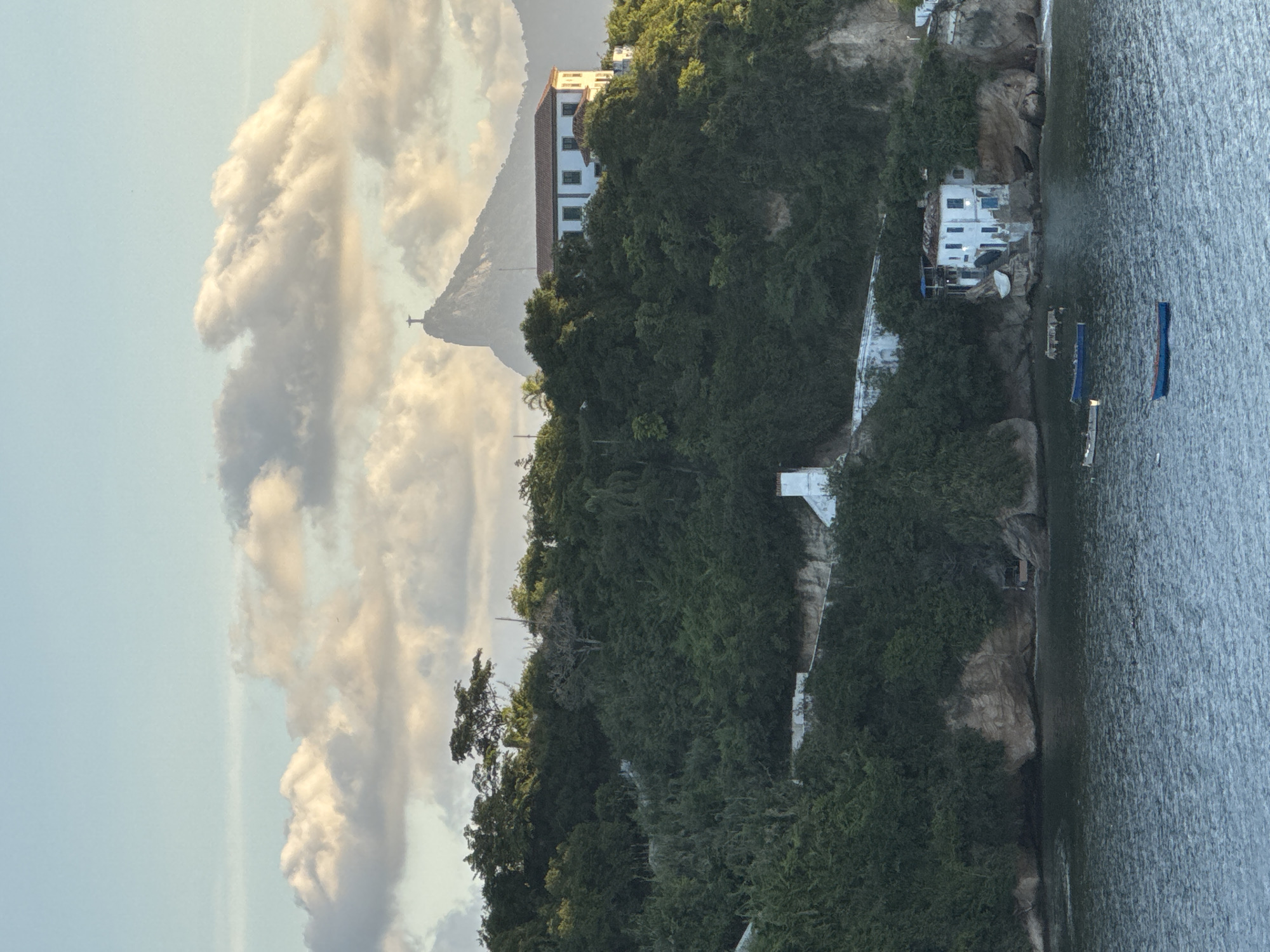 The image captures a view from the Guanabara Bay looking towards Niterói, Brazil. In the distance, the iconic Christ the Redeemer statue stands atop Corcovado Mountain, partially obscured by clouds. Closer to the foreground, a lush, green hillside is dotted with buildings. One prominent white building with a red tiled roof sits high on the hill, while smaller white structures are nestled closer to the water's edge. The water is calm, with several small boats visible. The scene is bathed in the warm, soft light of what appears to be either sunrise or sunset, casting a serene atmosphere over the landscape.