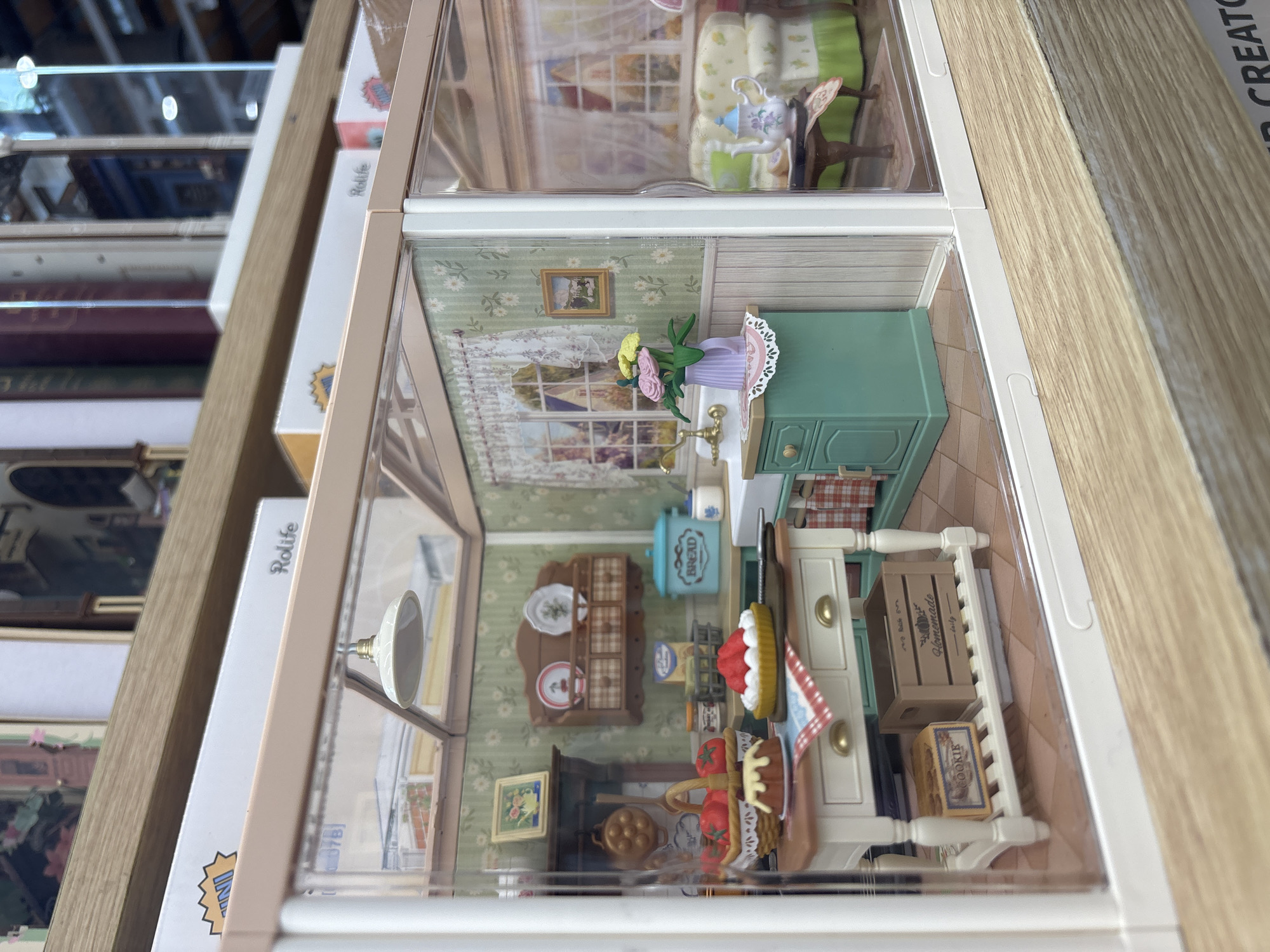The image shows a dollhouse display on a wooden shelf. The dollhouse is divided into two rooms. The bottom room appears to be a kitchen with miniature furniture, including a table, cabinets, a stove, and a sink. Food items like bread and cakes are also present. The top room contains furniture and decorations. The name "Rolife" is visible on the side of the dollhouse. The location is not inferable from the image content. The image was taken at a store.