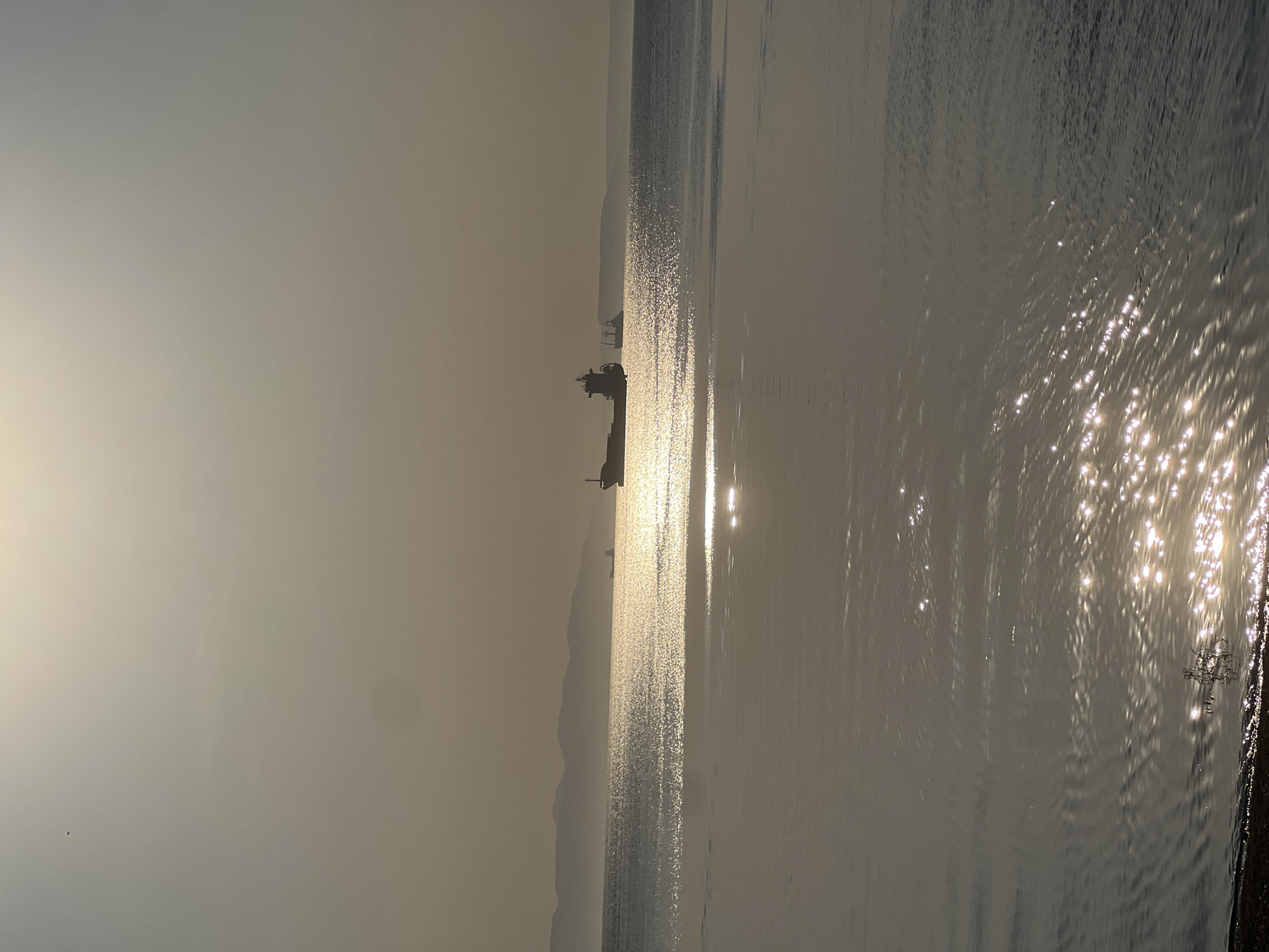 The image depicts a seascape in a hazy, light-filled environment, likely taken at dawn or dusk. The location is Vladivostok, Russia, as indicated by the presence of large cargo ships visible on the horizon. The sun's reflection creates a shimmering path across the water's surface, with the brightest reflections near the right edge of the frame where the water meets the shore. The water appears calm, with subtle ripples reflecting the sunlight. A large ship is visible on the horizon. The sky is obscured by haze or fog, creating a muted color palette. The coastline is dimly visible behind the ships.