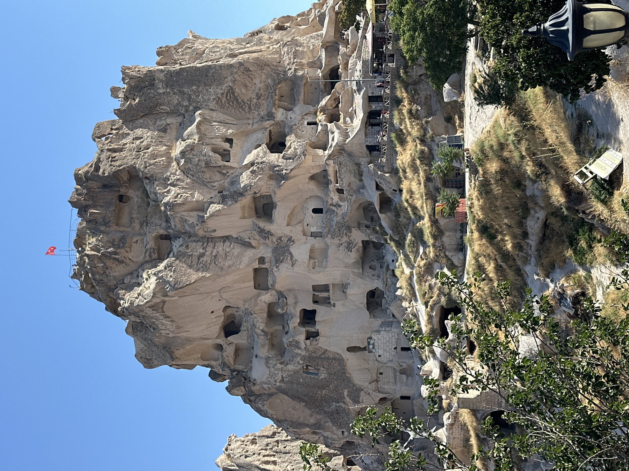 The image displays Uçhisar Castle, a prominent geological formation with numerous cave dwellings and carved structures, located in Uçhisar Beldesi, Turkey. The castle is a large, imposing rock outcropping against a clear blue sky. Many openings and windows are visible on its surface, indicating past human habitation. At the very top of the rock, a Turkish flag is prominently displayed on a flagpole.
Below the main rock formation, a pathway winds through dry, grassy terrain dotted with sparse trees and shrubs. On a lower level of the rock face, a row of businesses, possibly cafes or shops, are set up with tables and umbrellas, suggesting a tourist area. Several people can be seen in this area, some seated at tables and others milling about. In the foreground, a leafy green tree partially obscures the view of the rock face and its cave openings. A dark, ornate lamp post stands in the bottom right corner of the image. The overall scene depicts a historical and geological site with evidence of both ancient use and modern tourism.