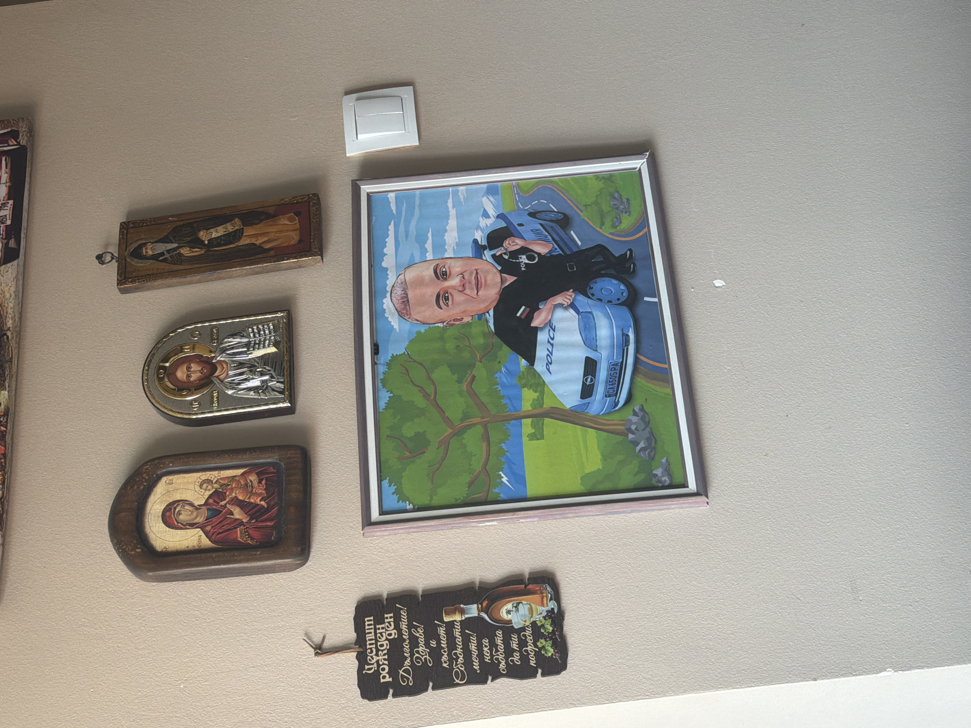 The image displays a section of a light-colored wall, likely in a home or office in Shumen, Bulgaria, adorned with various decorative items.
The central and largest item is a framed caricature drawing depicting a male figure, presumably a police officer, with a prominent head and light-colored hair. He is shown wearing a dark police uniform shirt, holding handcuffs, and leaning against a light blue police car. The car has "POLICE" written on its side and a visible Bulgarian license plate "CA 6505 PA" (indicating Sofia registration). The background of the caricature features a blue sky, green trees, and a winding road.
To the left of the caricature, a vertical arrangement of three religious icons is displayed. The uppermost is a rectangular, wooden-framed icon of a male saint. Below it is a silver-colored, arch-shaped icon featuring Christ. The lowest icon is a wooden-framed depiction of the Virgin Mary holding the Child Jesus.
Below the caricature, a dark, irregularly shaped wooden plaque hangs. It features an illustration of a bottle of spirits and a glass with grapes. Bulgarian text is inscribed on the plaque, wishing "Честит рожден ден" (Happy Birthday), "Дълголетие!" (Longevity!), "Здраве!" (Health!), "и късмет!" (and luck!), "Сбъднати мечти!" (Fulfilled dreams!), and "нека съдбата да ти подреди" (may fate arrange for you...).
A white rectangular light switch is mounted on the wall above the caricature. No specific activity is occurring; the scene captures a static arrangement of personal decorations. The indoor setting provides no specific cues about the time of day or weather.