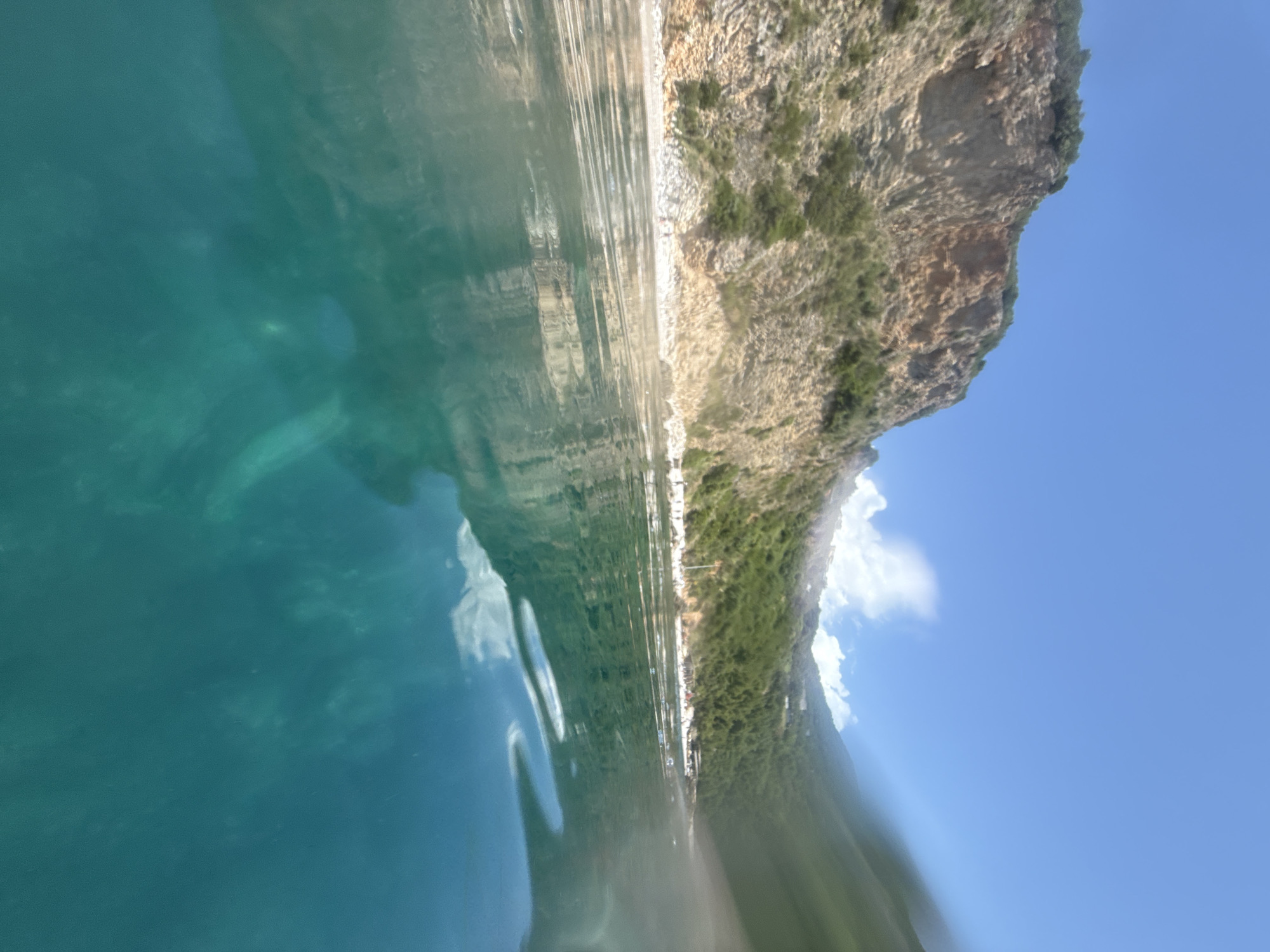 The image is a split-level photograph, capturing both an above-water and a below-water scene under bright daylight.
Above the distinct waterline, a steep, rocky cliff face dominates the left and center, exhibiting various shades of brown, tan, and grey. Sparse green vegetation, consisting of bushes and trees, clings to the cliff, becoming denser towards the upper and right portions of the land. At the base of the cliff, a narrow, light-colored pebble or rocky beach is visible, populated by several small, indistinct human figures engaged in leisure activities such as sunbathing or walking. On the far right, additional green hills covered in vegetation extend, with some distant structures potentially indicating human habitation or facilities. The sky is clear and blue, with scattered white cumulus clouds visible towards the upper right, partially obscured by a blurred effect or water splash on the right side of the frame. The water's surface exhibits ripples and reflections of the cliff face and sky.
Below the waterline, the water is transparent with a distinct turquoise-green hue. Indistinct, elongated dark shapes are visible submerged in the deeper parts of the water, particularly towards the lower left and center-right, suggesting underwater topography or objects. The visibility below the surface is high, allowing for clear observation of the water's color and the shadowy forms within. The overall scene depicts a sunny coastal environment in Unknown, Unknown.